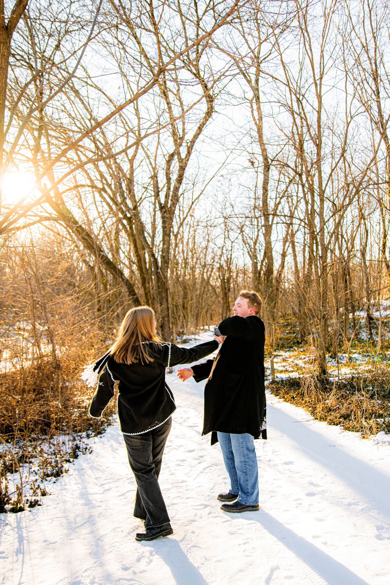 Couple holding hands walking on a snowy winter path surrounded by bare trees with golden sunset light.