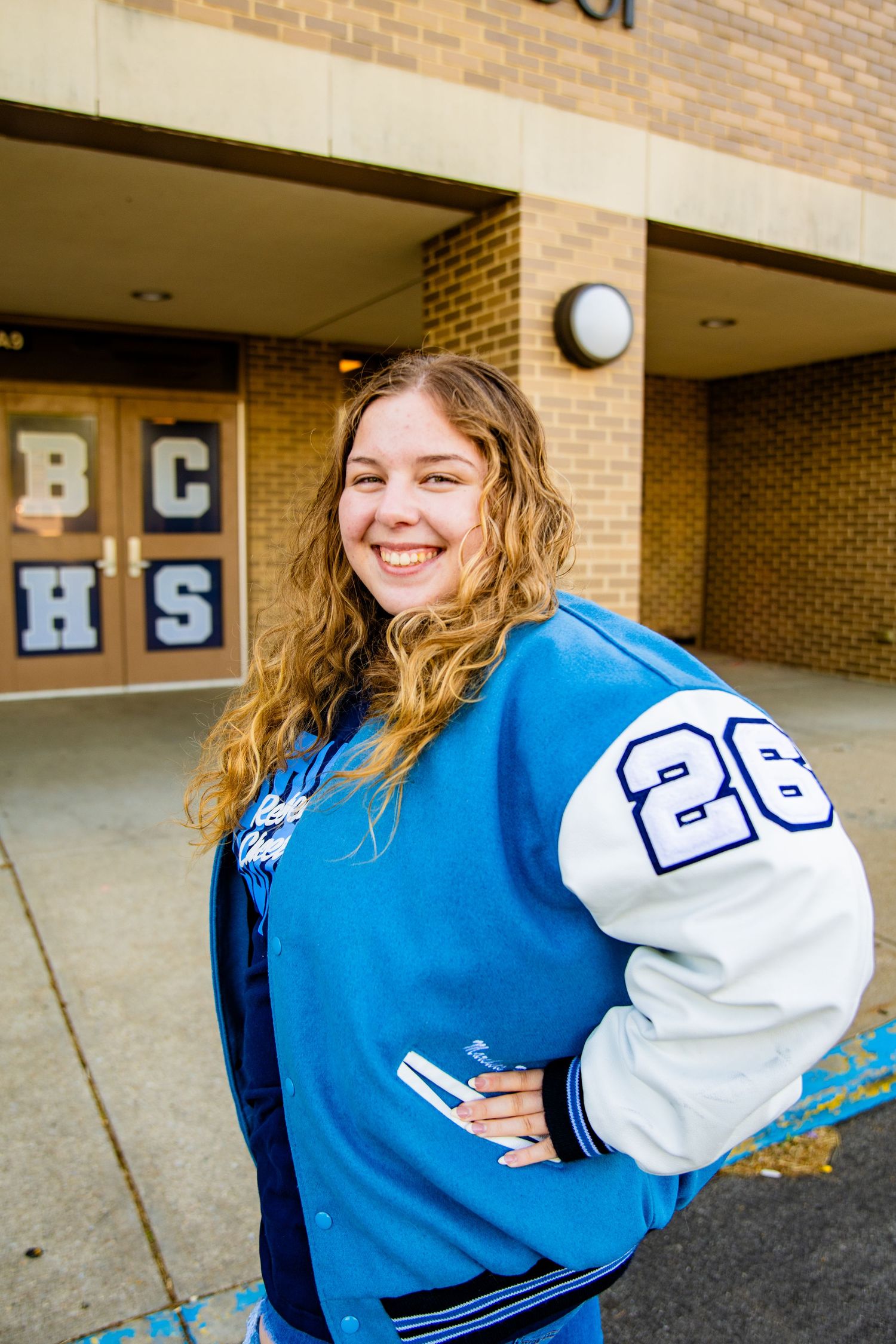 Smiling teenage girl in blue and white BCHS letterman jacket standing outside brick high school building.