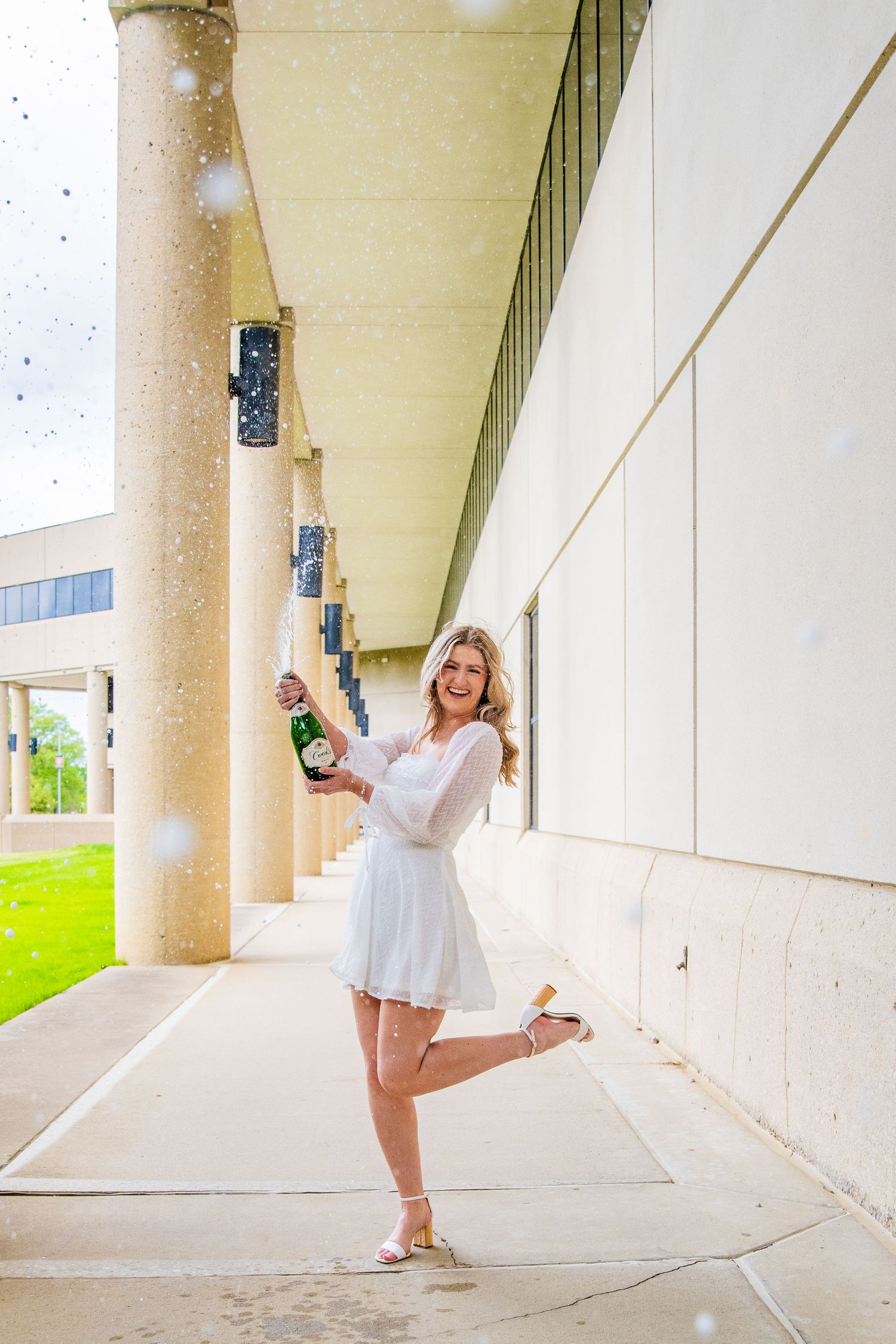 Young woman in white dress joyfully popping champagne bottle outdoors near building columns.