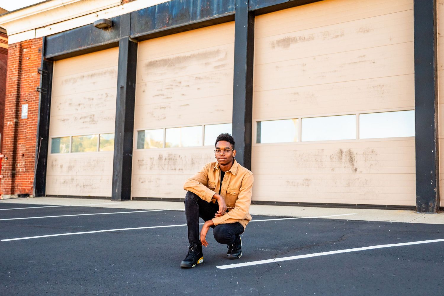 Young man in yellow jacket crouching in parking lot in front of industrial garage doors.