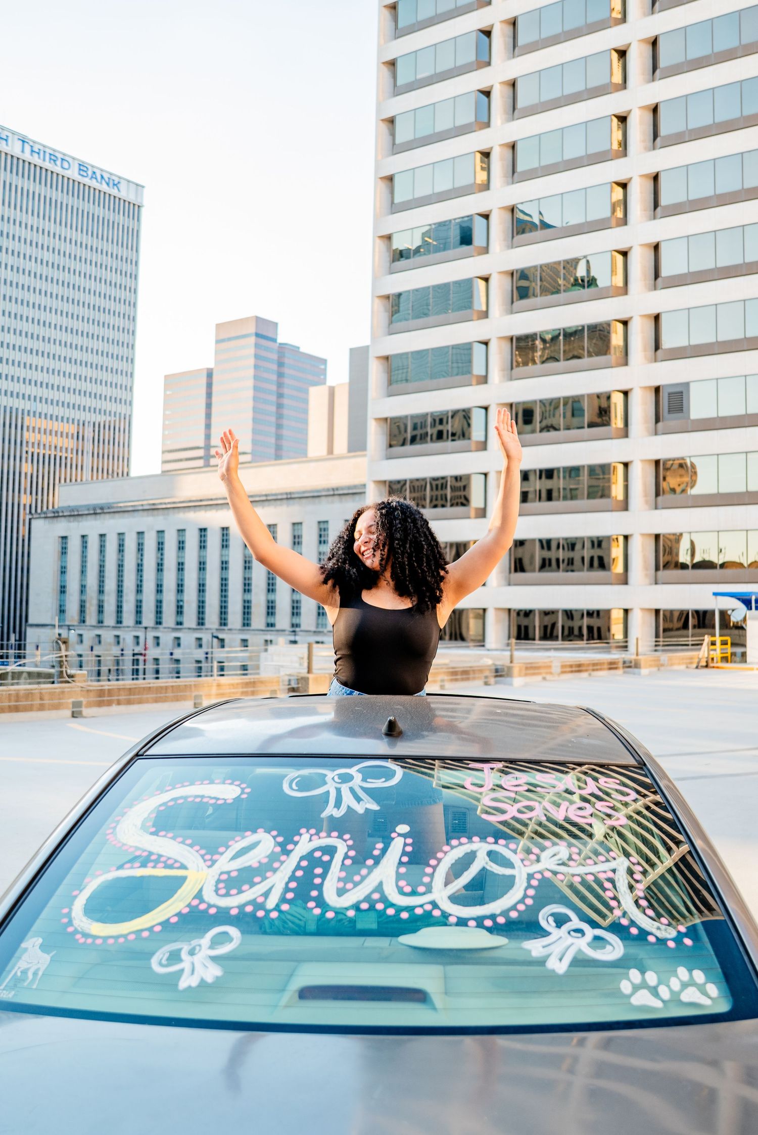 A senior student celebrates through a sunroof with arms raised, car window painted with Senior, city skyline behind.