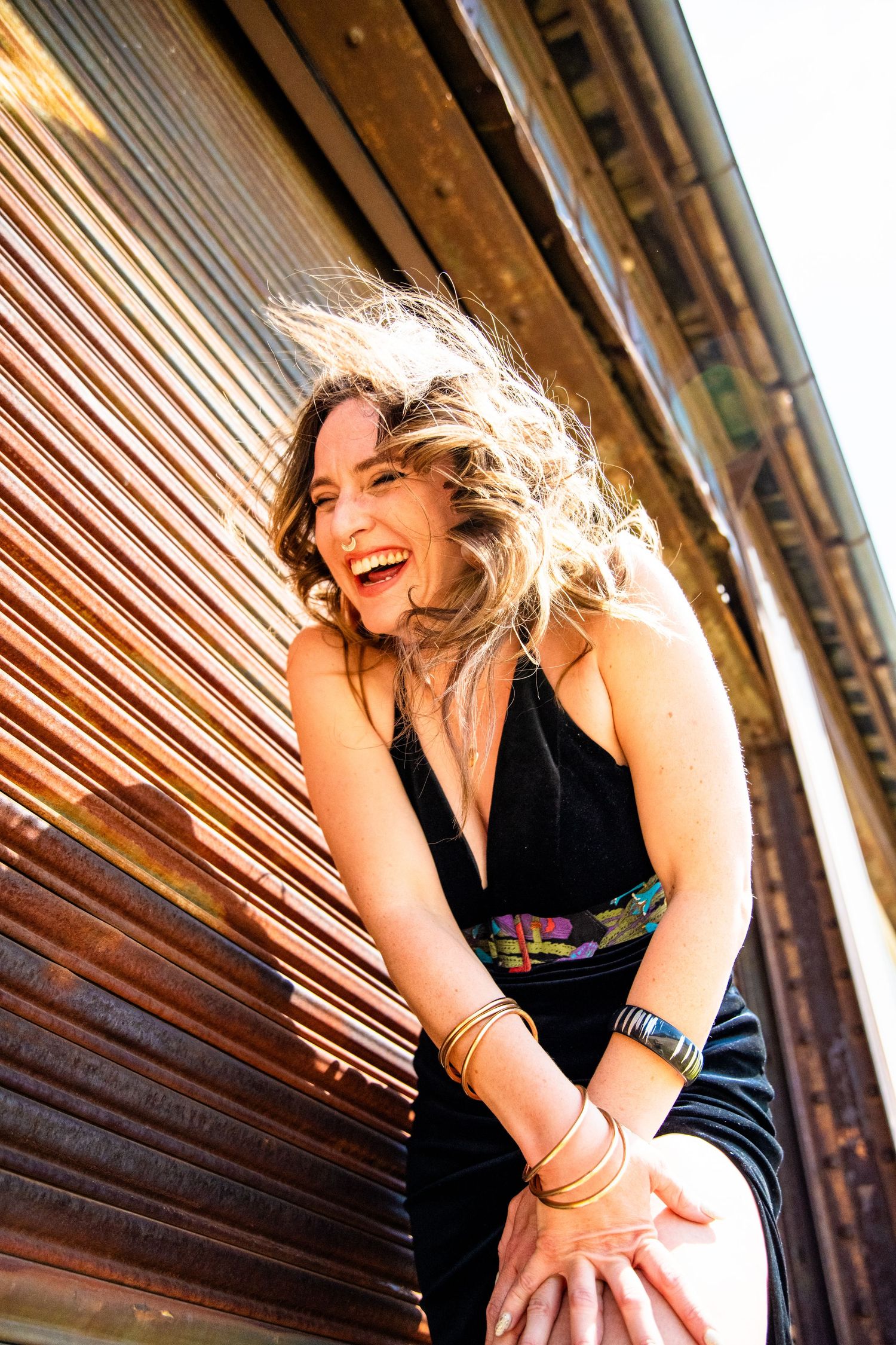 Laughing woman in black halter dress with gold bangles leaning against rustic corrugated metal wall outdoors.
