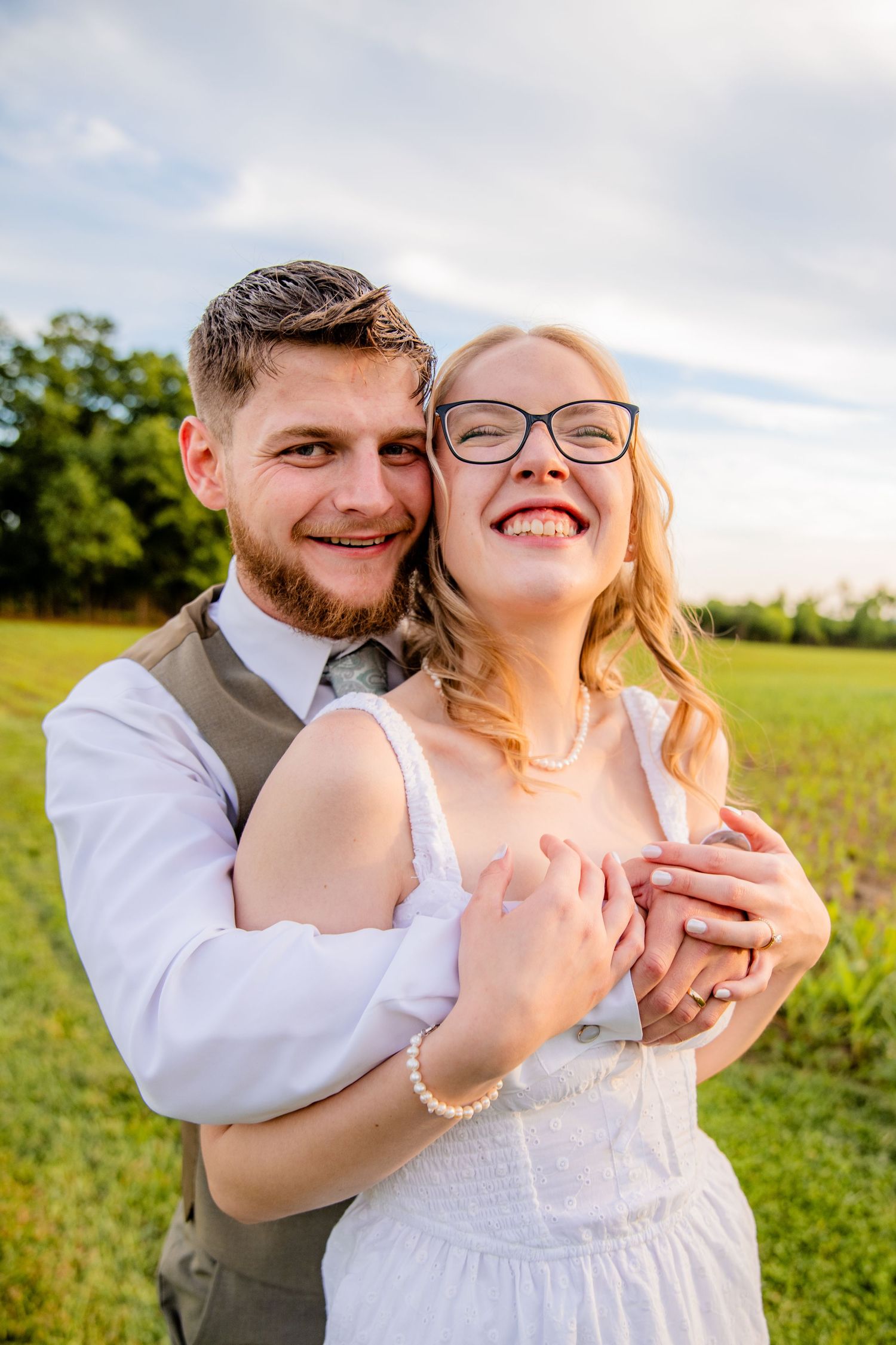 Joyful bride in white dress and groom in vest embracing in a sunny green countryside field.