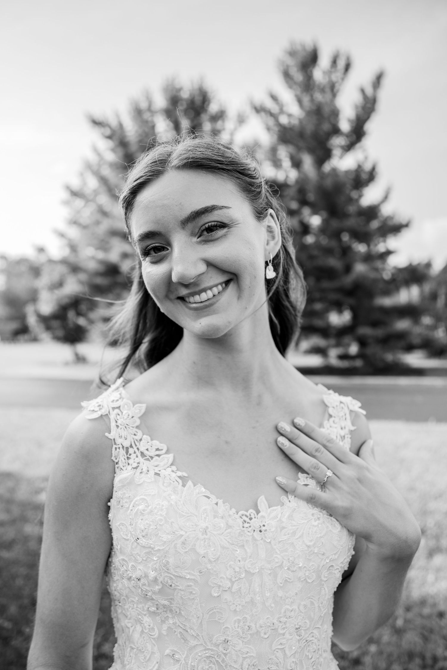 A smiling bride in a lace wedding gown poses outdoors in a black and white portrait photograph.