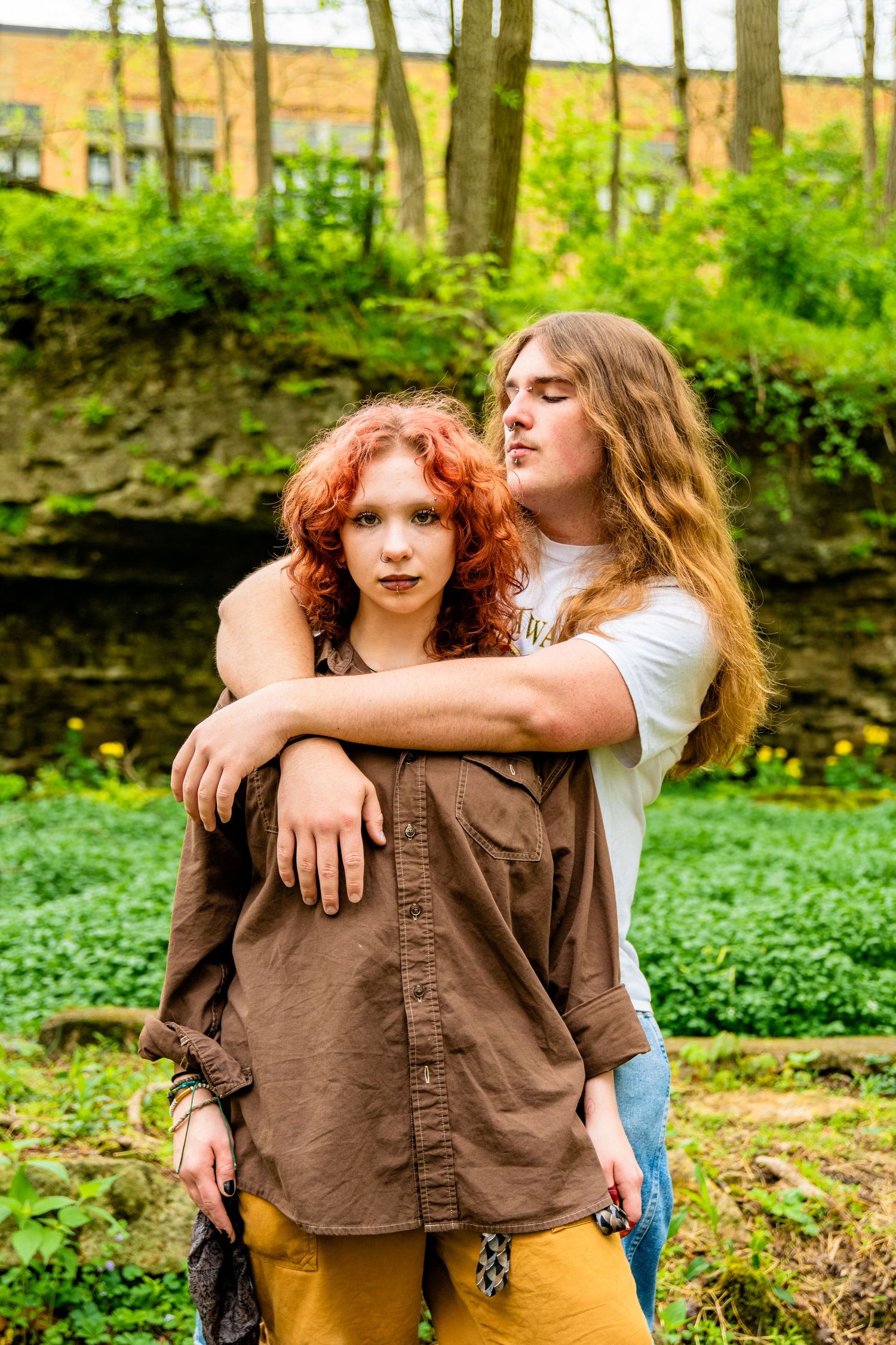 A young couple poses outdoors, woman with red curly hair in brown shirt, man with long hair embracing her from behind.