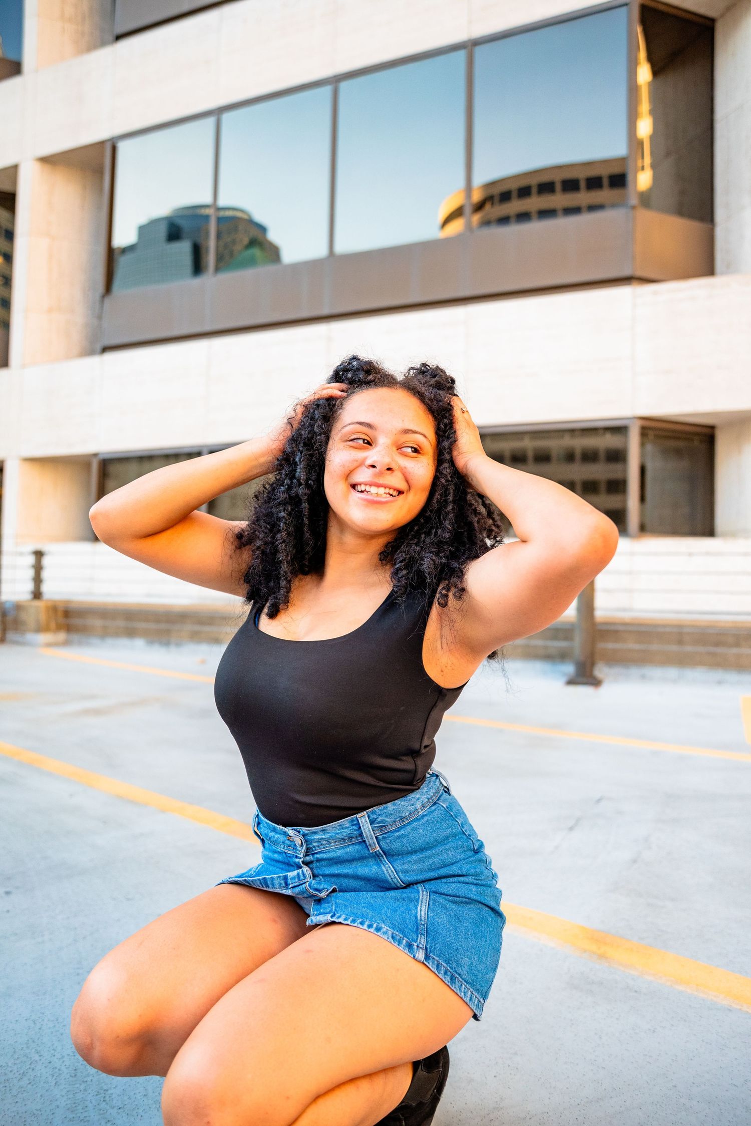 Smiling young woman with curly hair in black tank top and denim skirt posing outdoors near urban building.