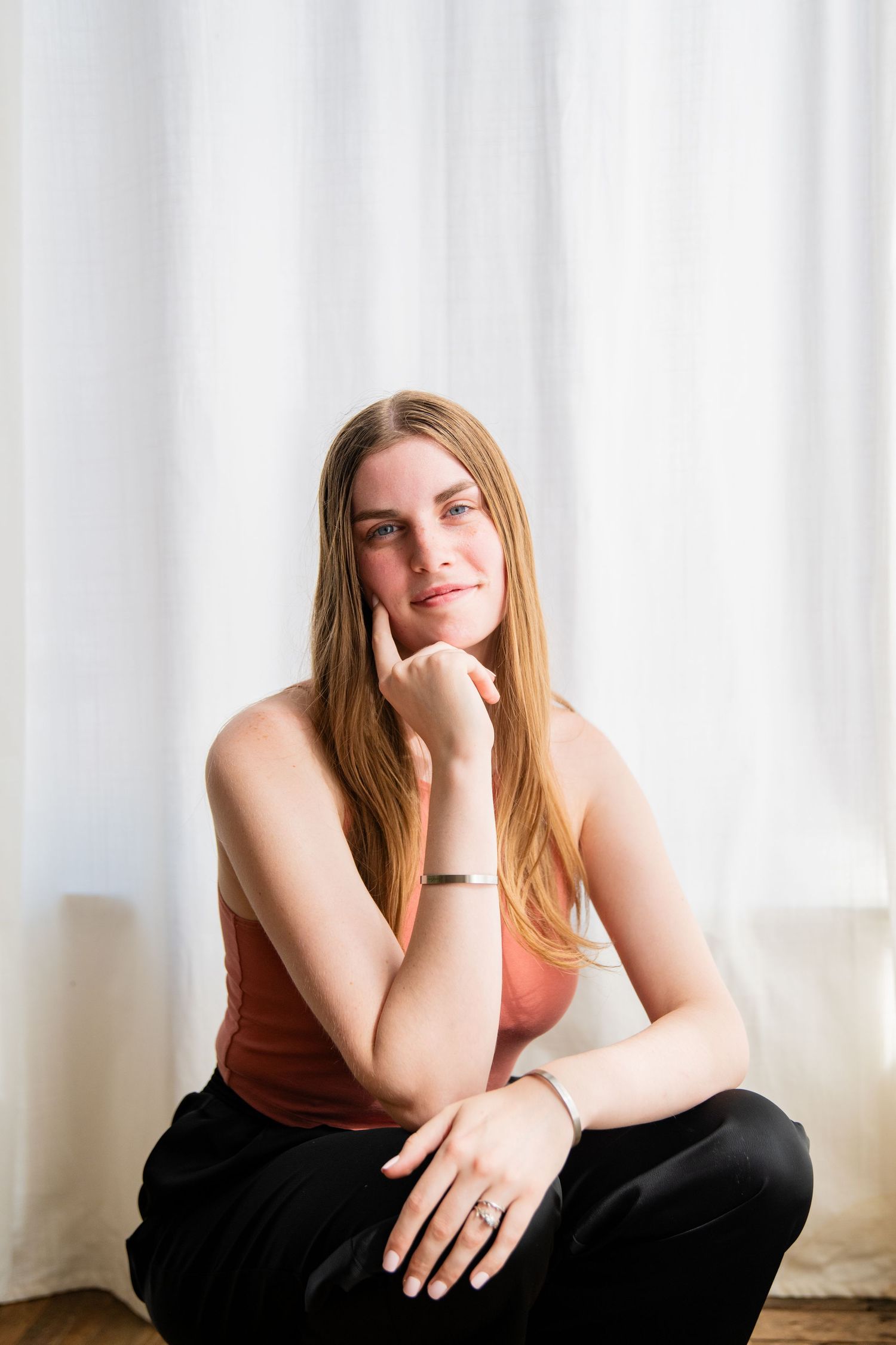 Young woman with long blonde hair sitting cross-legged, wearing a pink tank top and black pants, resting chin on hand.