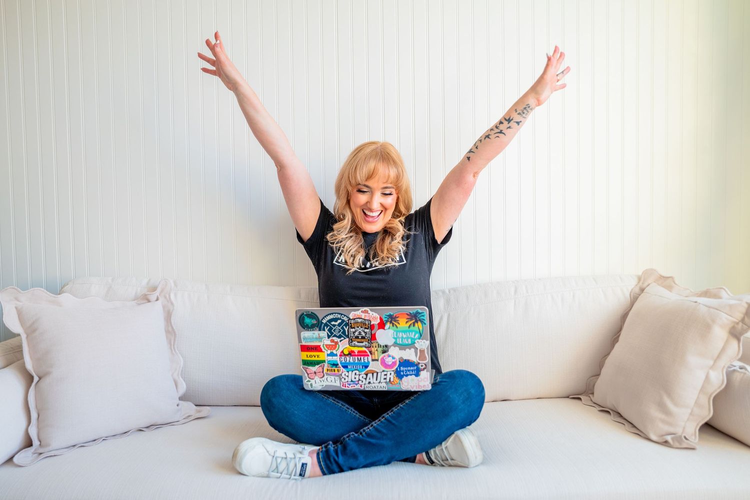 Excited blonde woman with tattoo raises arms in victory while using sticker-covered laptop on white couch.