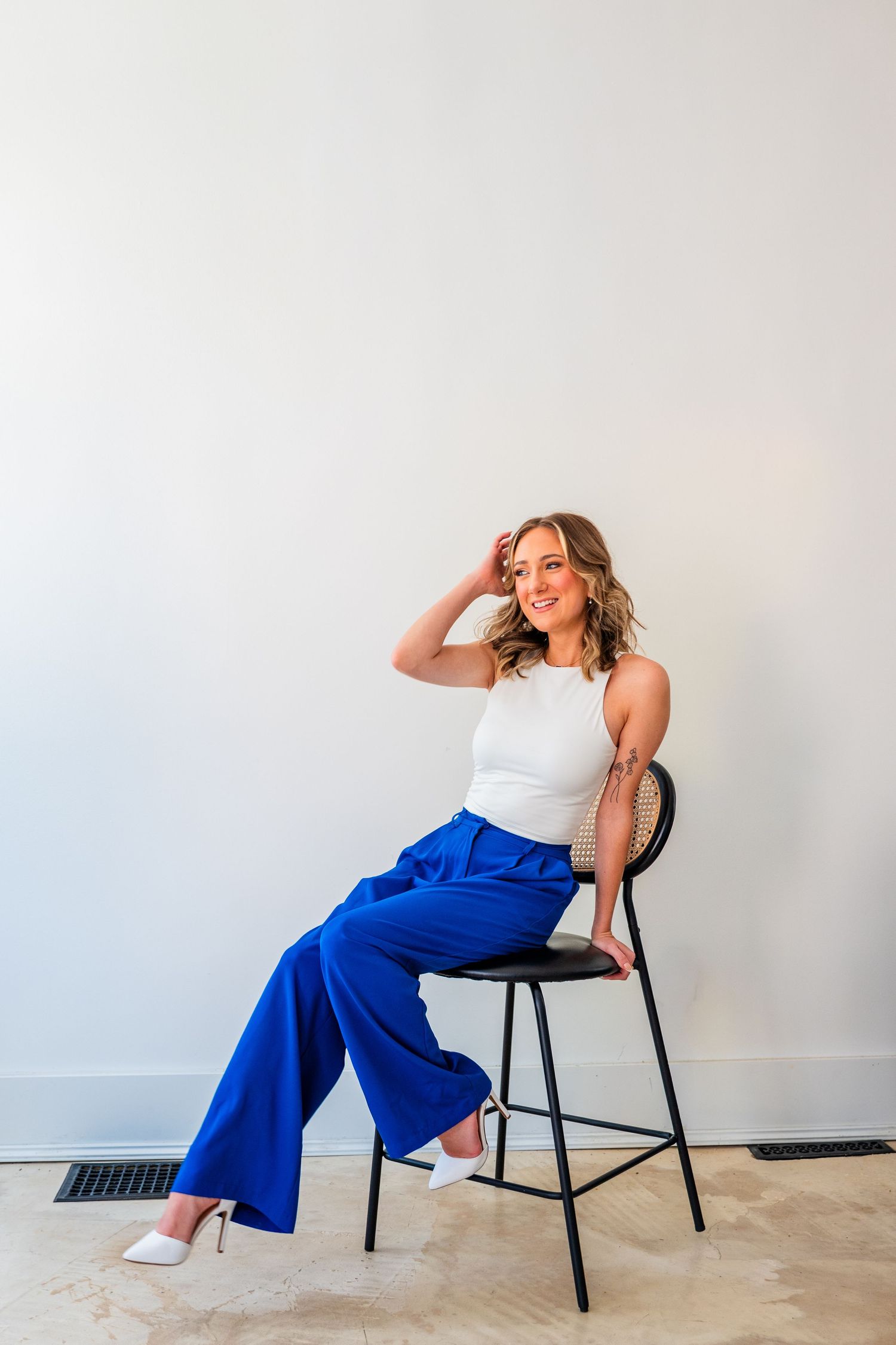 Woman in white sleeveless top and royal blue wide-leg pants sits on black chair, smiling against white wall.