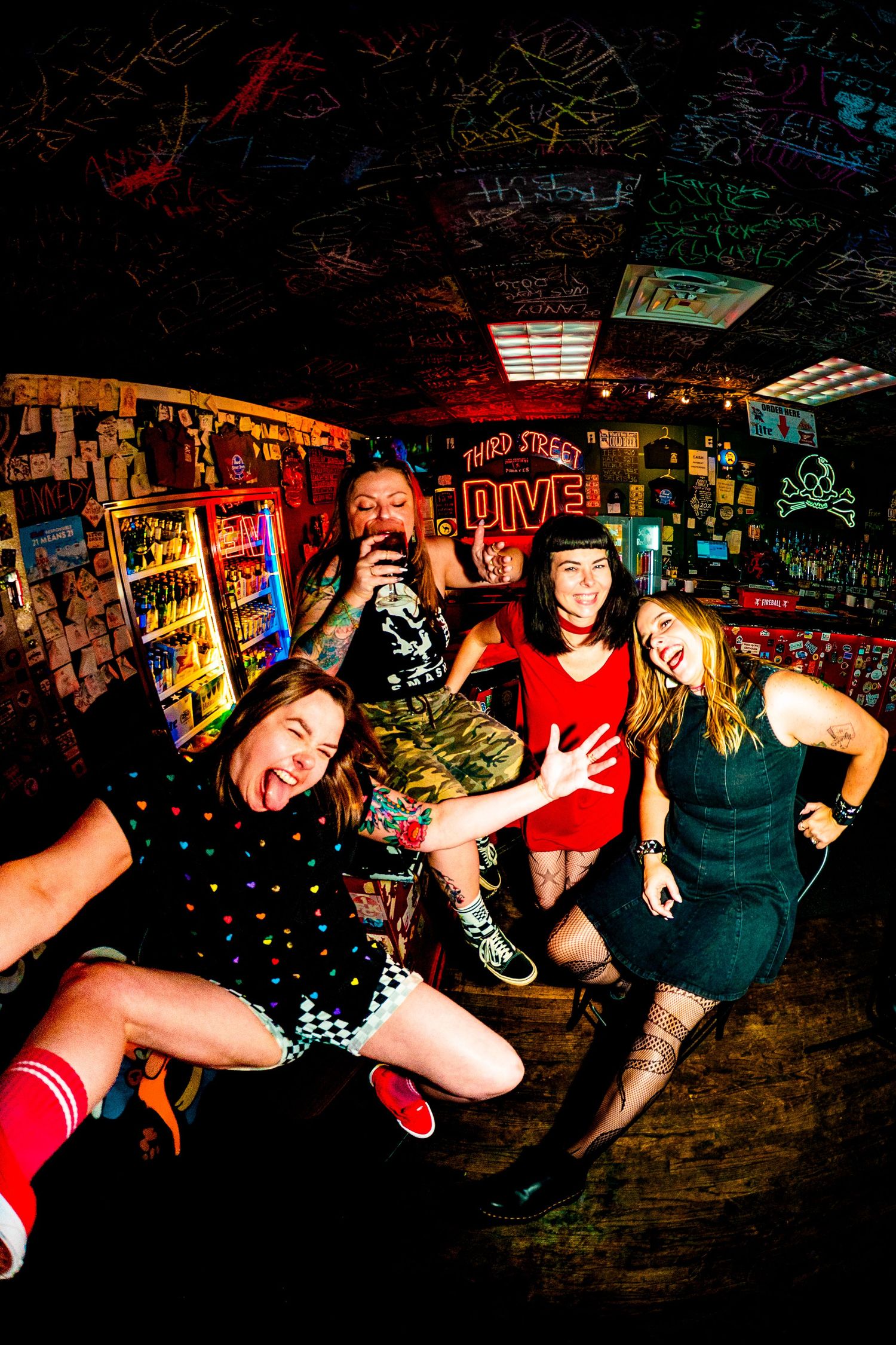Four friends pose energetically at Third Street Dive bar, a colorful graffiti-covered venue with neon signs.