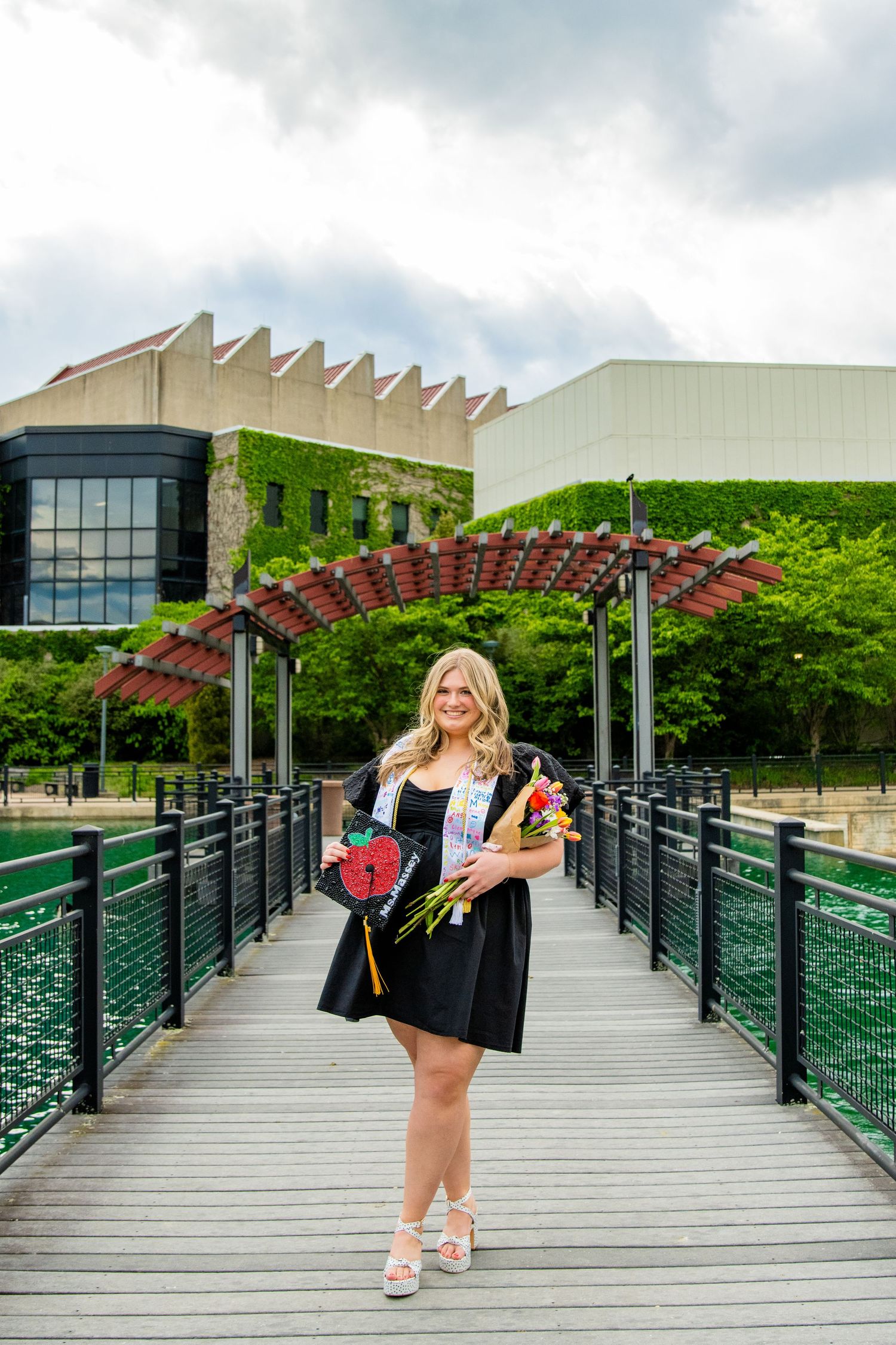 Young woman in graduation attire holding flowers and cap on a bridge, celebrating her academic achievement outdoors.