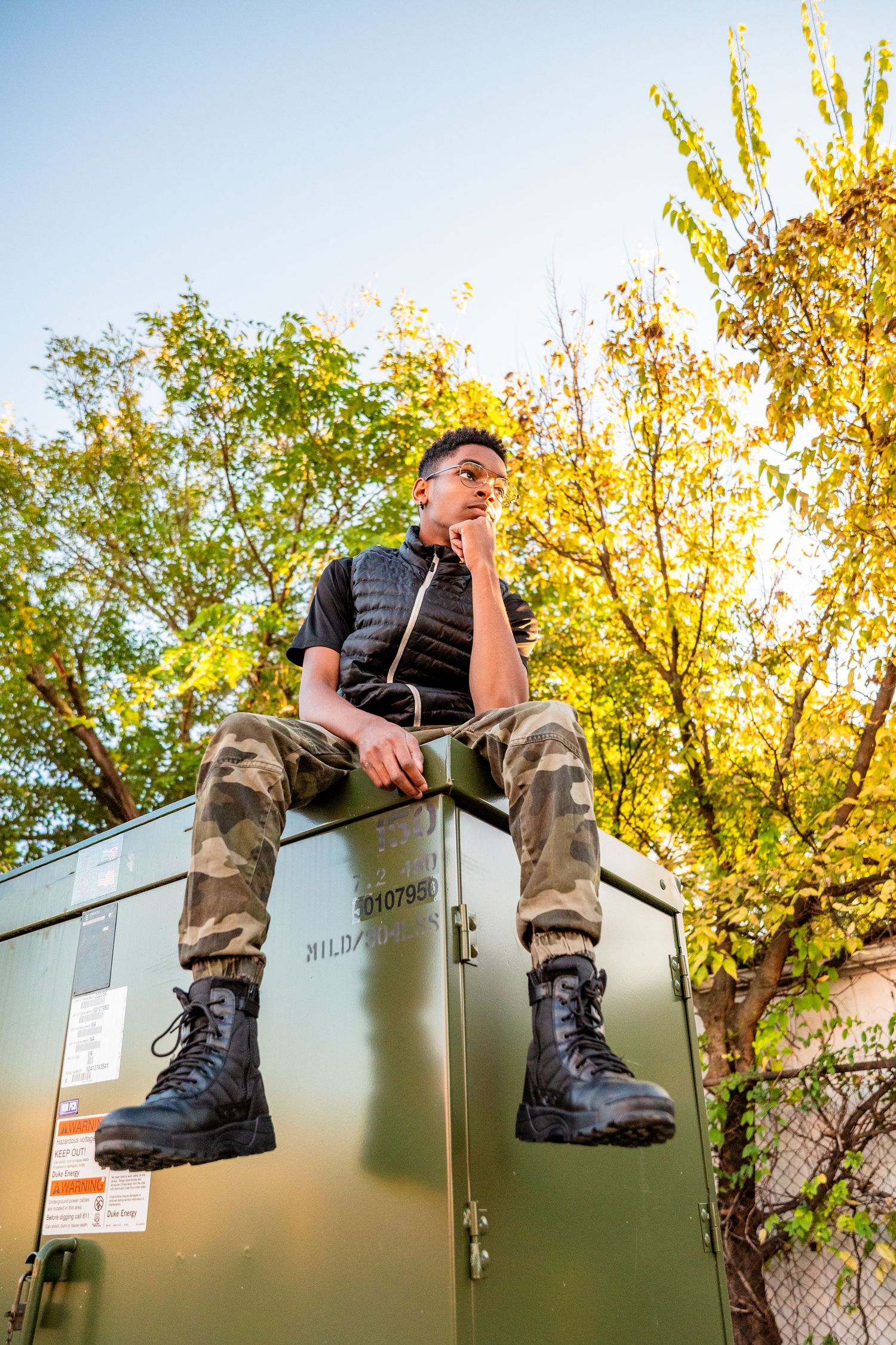 Young man in camo pants and combat boots sitting atop a green metal box outdoors with autumn trees behind him.