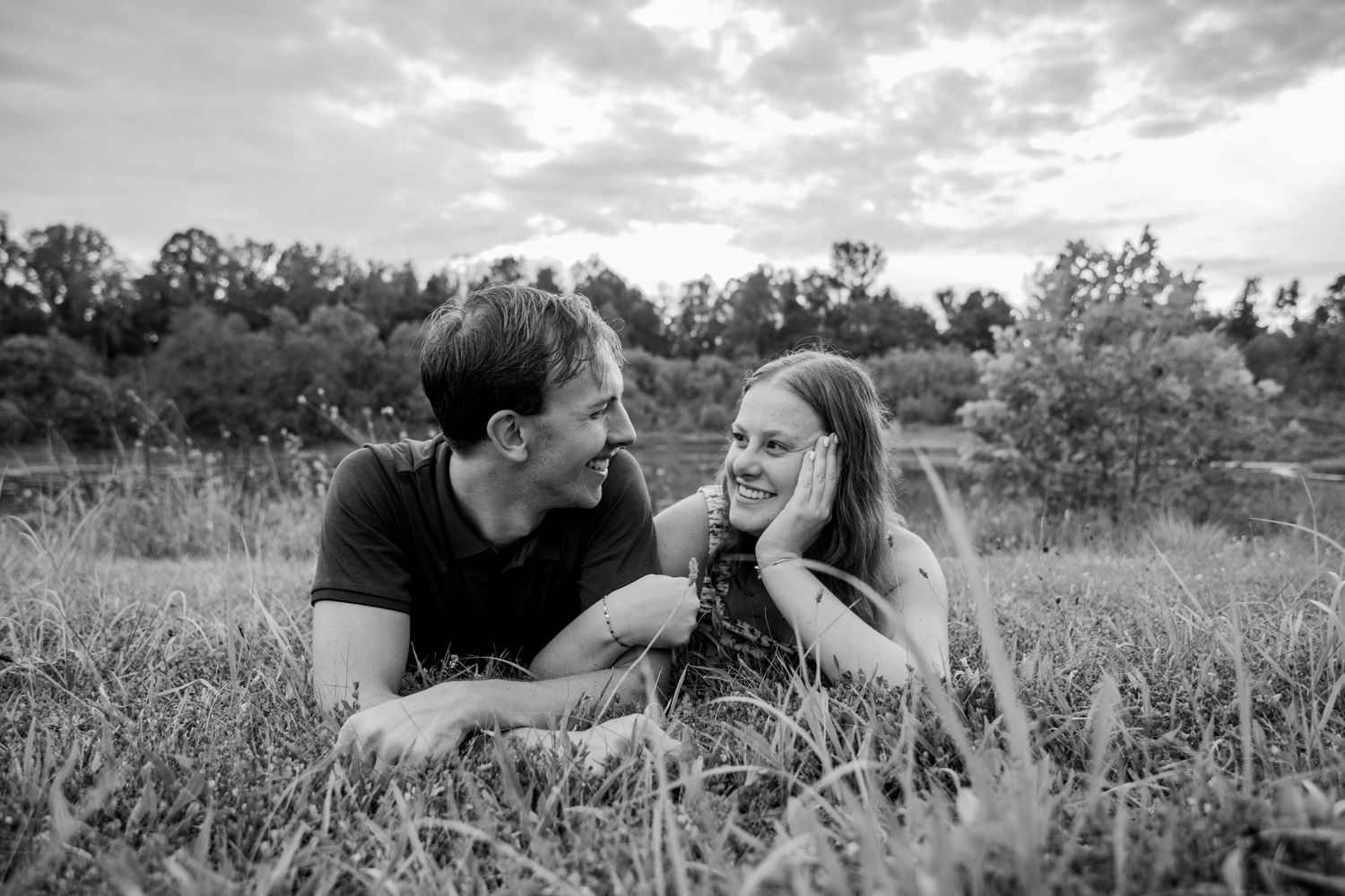 Black and white photo of a couple lying in grass, smiling at each other with trees in the background.