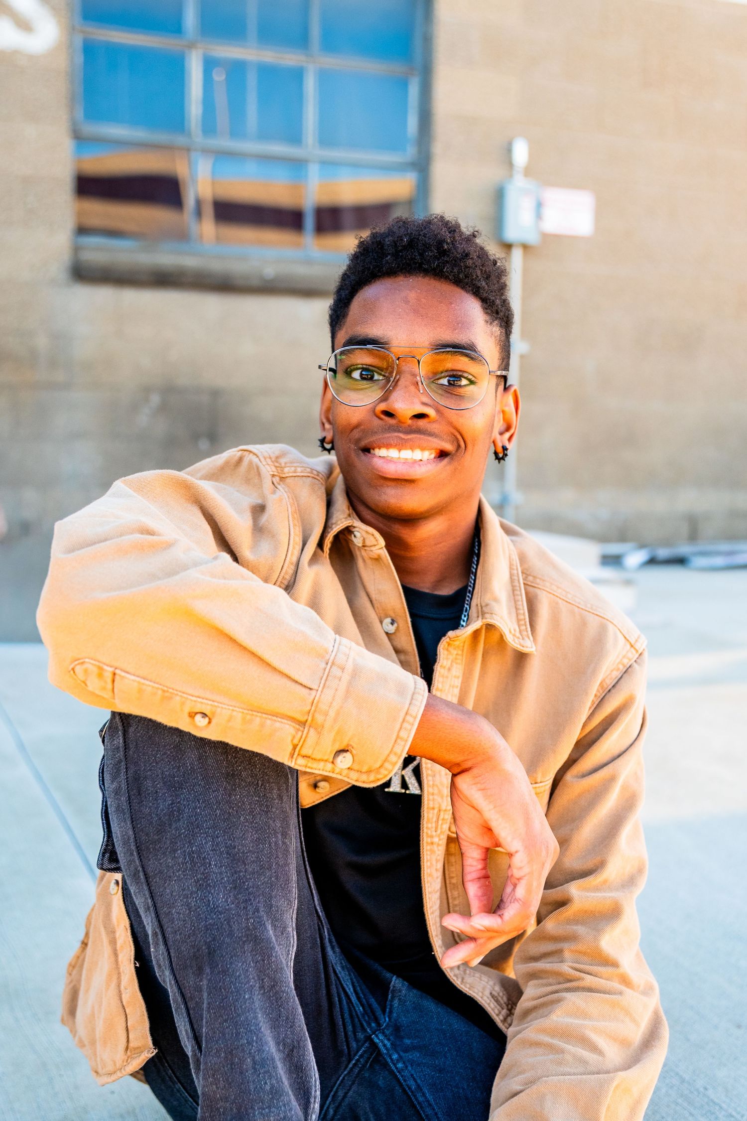 Smiling young Black man in tan denim jacket and glasses sits outdoors, posing confidently against a brick building.