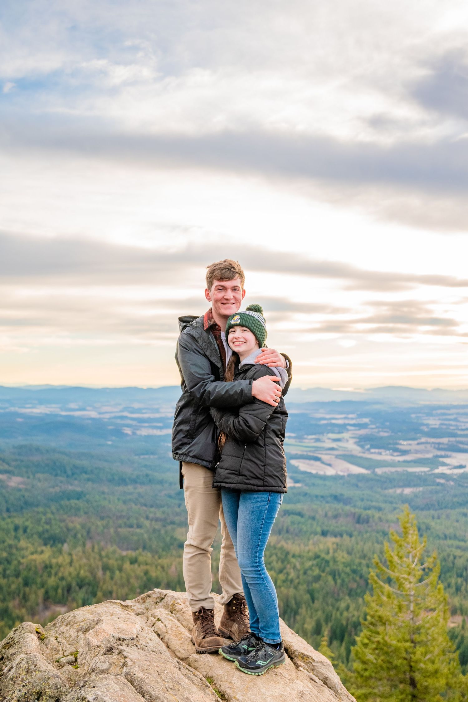 Couple embracing on a rocky mountain summit with panoramic forest and valley views under a cloudy sky.