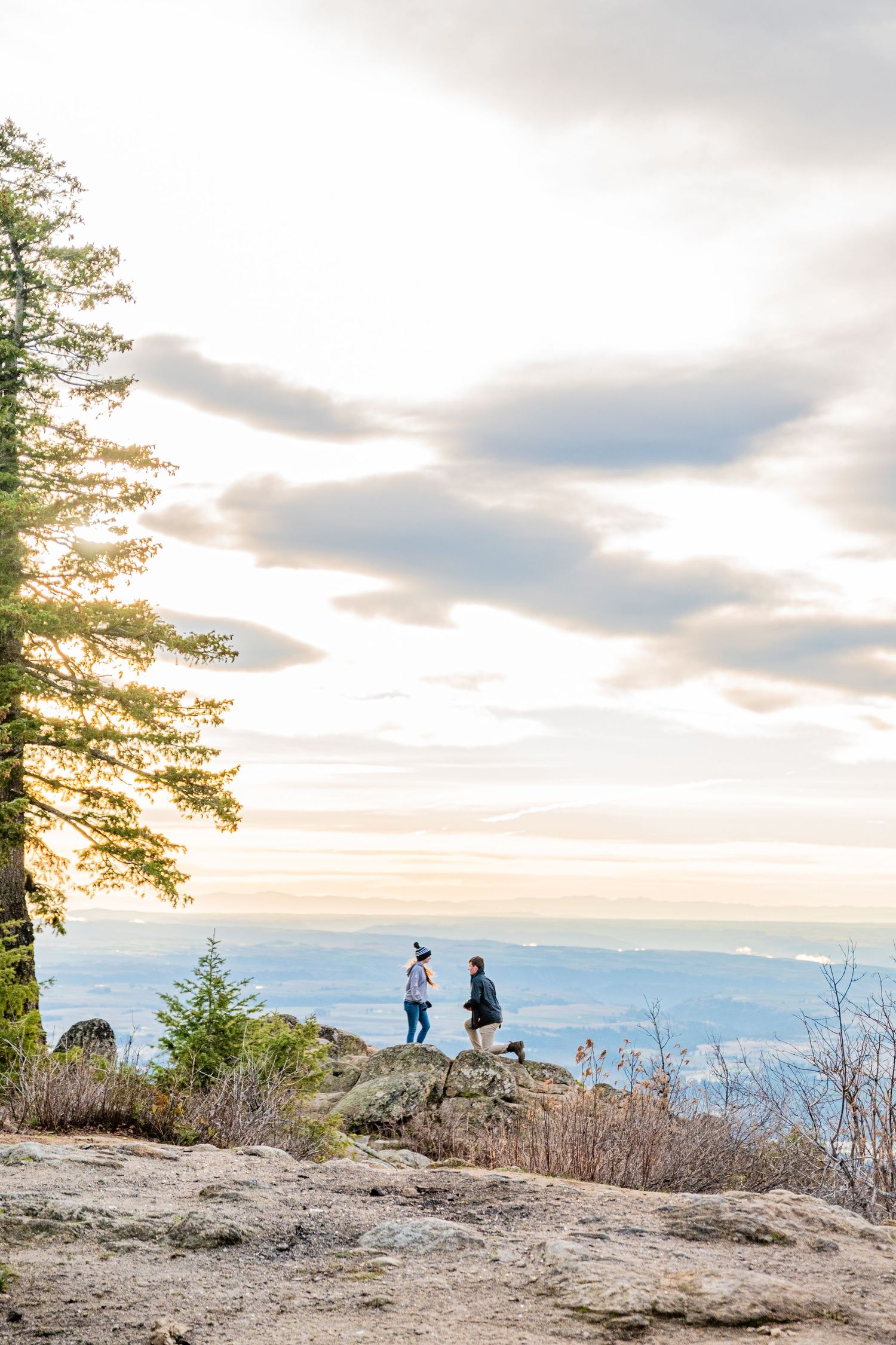 Two hikers stand on a rocky mountain summit overlooking a vast valley landscape at golden hour sunset.