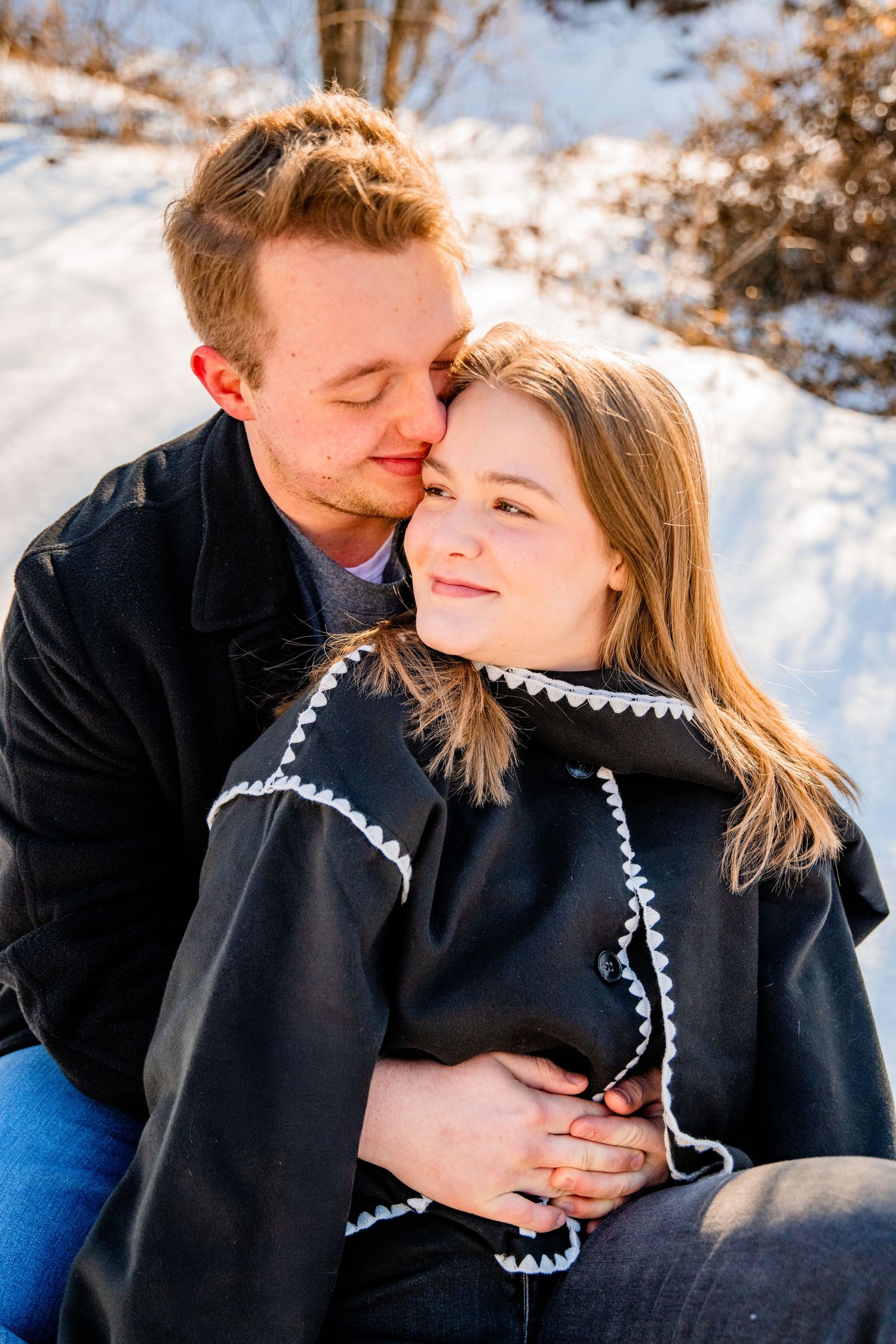 Couple embracing in snowy winter setting, woman smiling in black cape coat, man nuzzling her cheek affectionately.