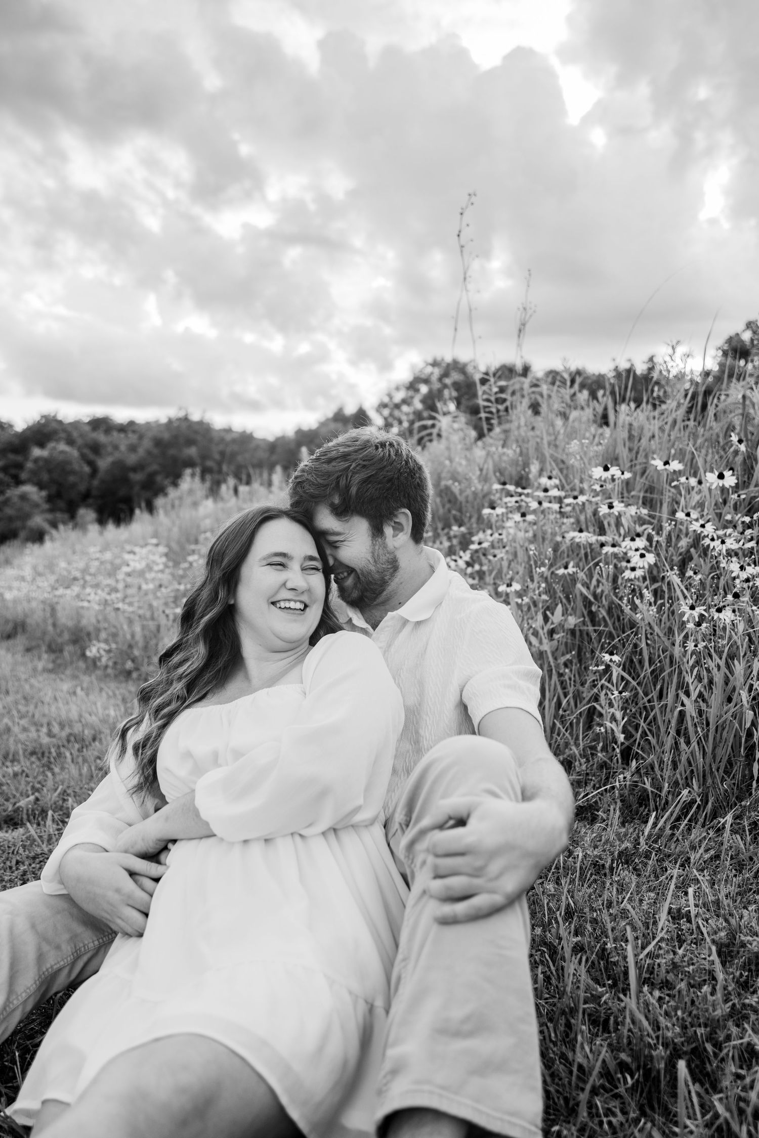 Black and white photo of a couple sitting in a wildflower field, laughing and embracing under a cloudy sky.