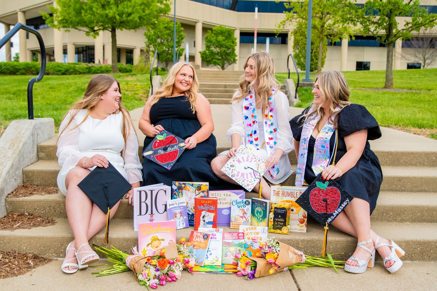 Four female graduates in black dresses sit on steps with caps, flowers, and colorful gifts celebrating their graduation day.