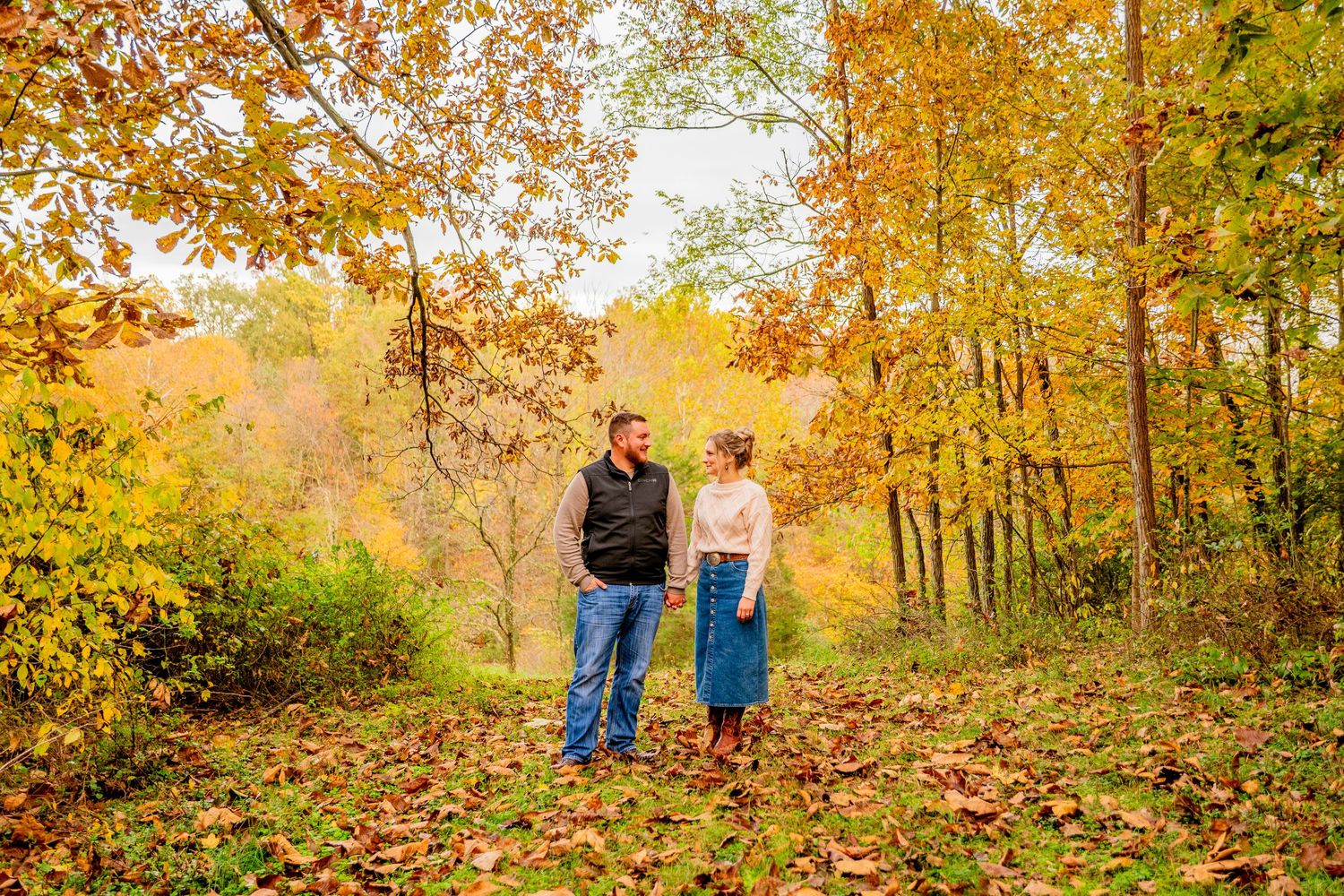 Couple holding hands in autumn forest with colorful fall foliage in orange, yellow, and green hues.