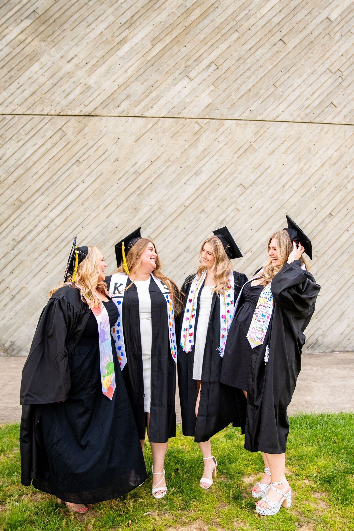 Four female graduates in black caps and gowns with colorful stoles celebrate together outdoors.