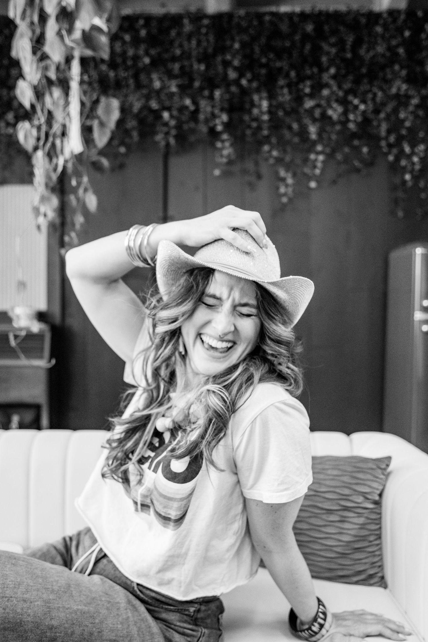 A laughing woman in a cowboy hat and white t-shirt sits on a couch in a black and white photo.