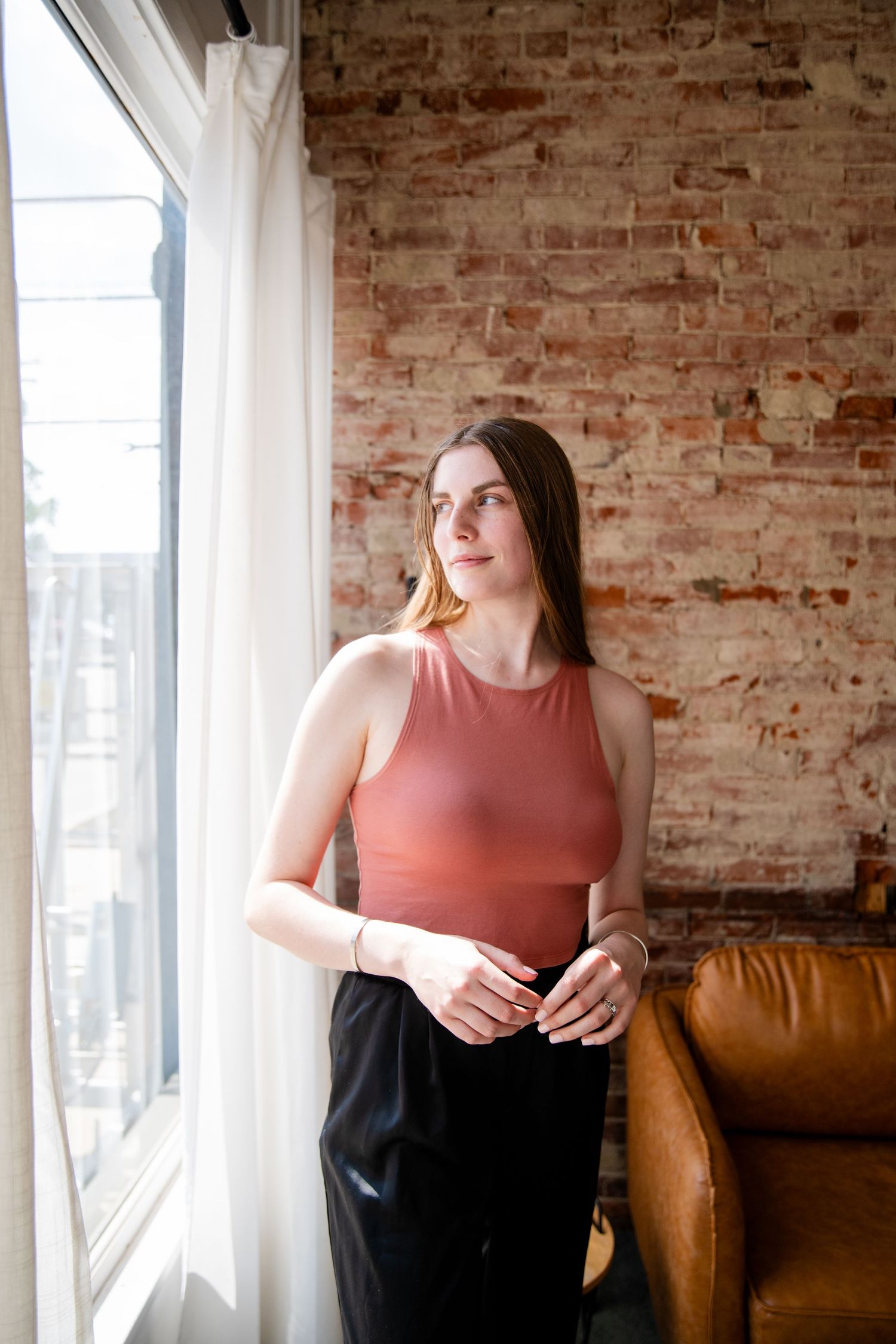 Young woman in pink tank top and black pants standing by a sunlit window with exposed brick wall and leather chair.