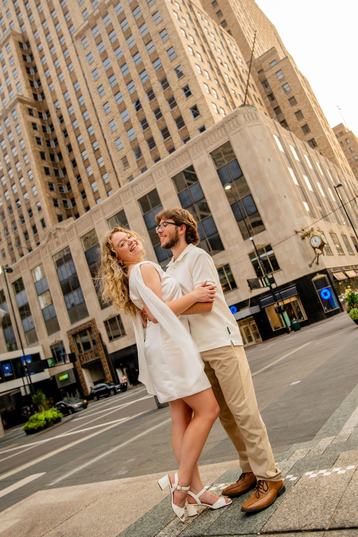 Couple embracing on urban street corner with tall beige skyscraper backdrop, woman in white dress, man in khaki pants.