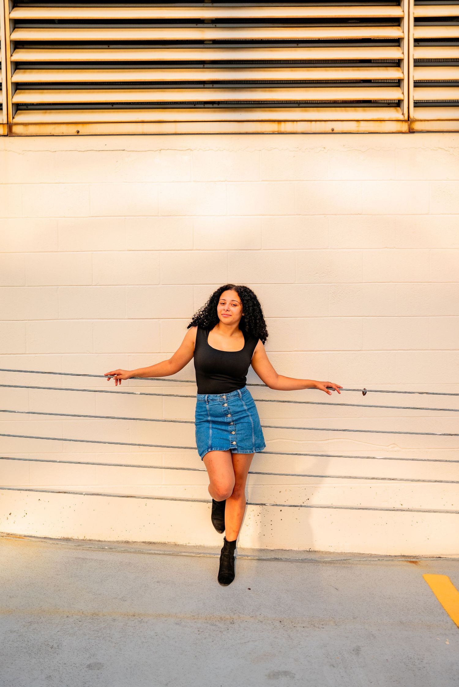 Young woman in black top and denim skirt posing against a white wall in golden hour light.