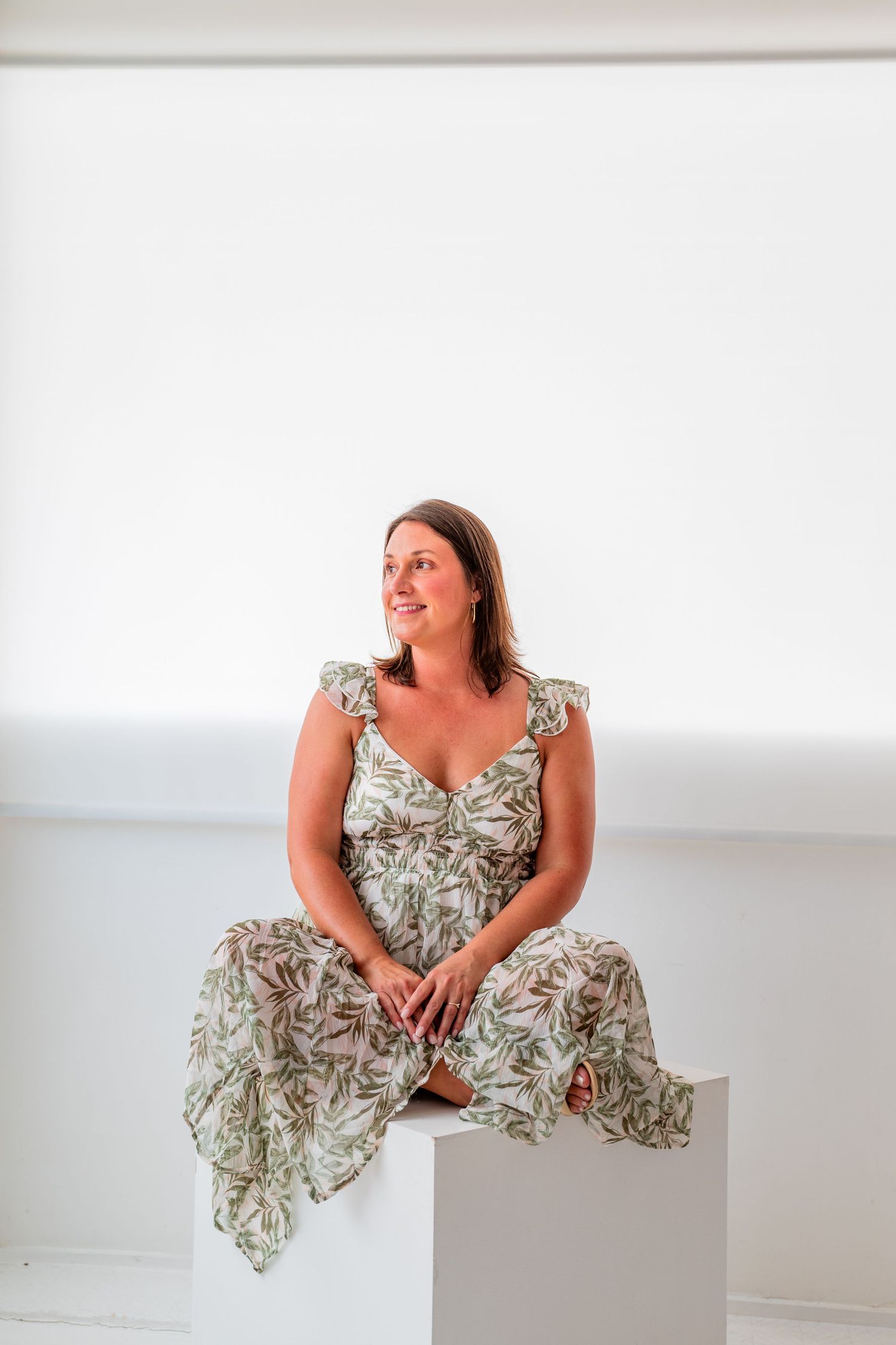 Woman in floral green dress sitting on white cube, smiling and looking to the side in bright white studio.