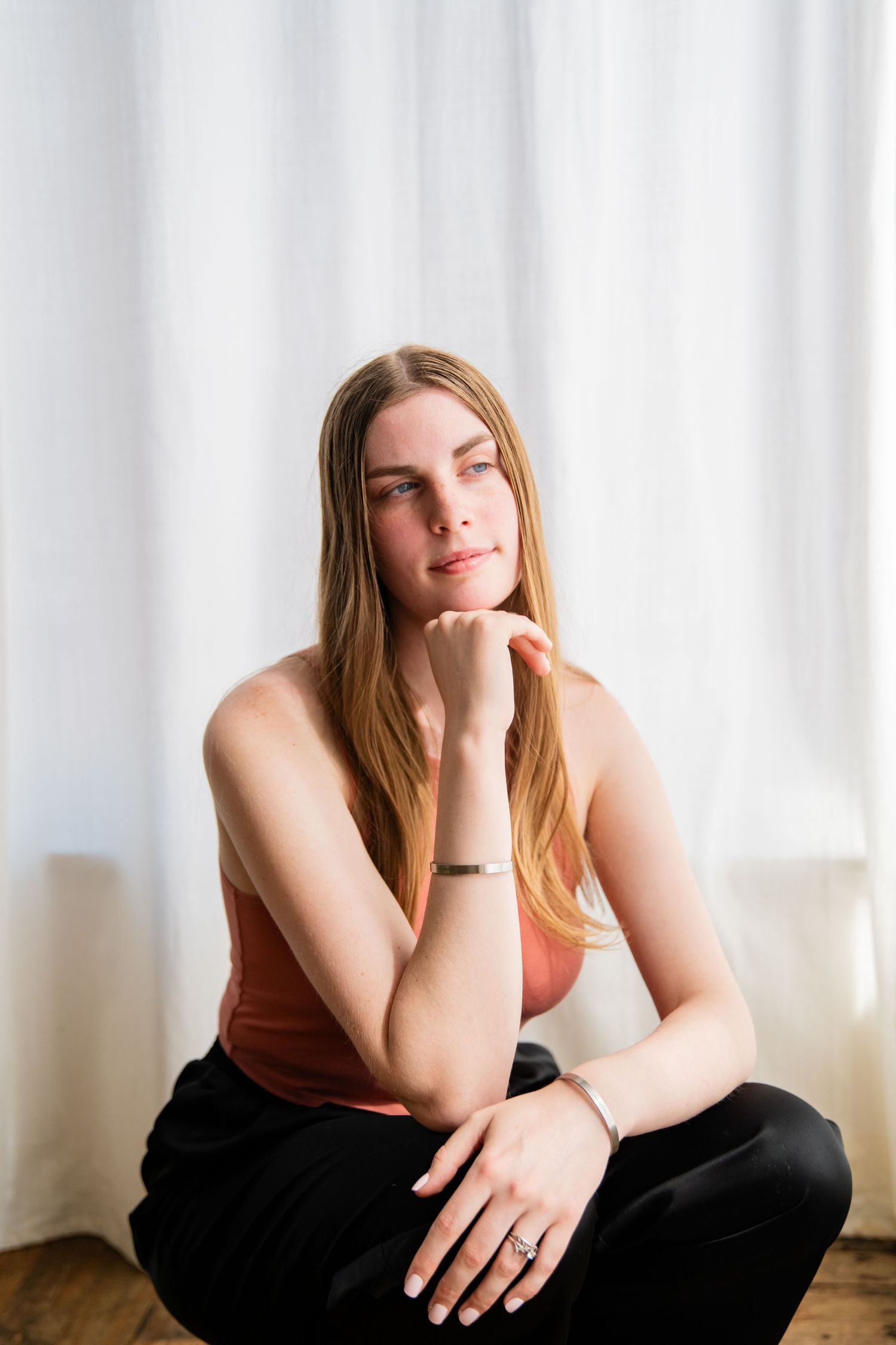 Young woman with long blonde hair sits cross-legged in a coral tank top and black pants, resting chin on hand thoughtfully.