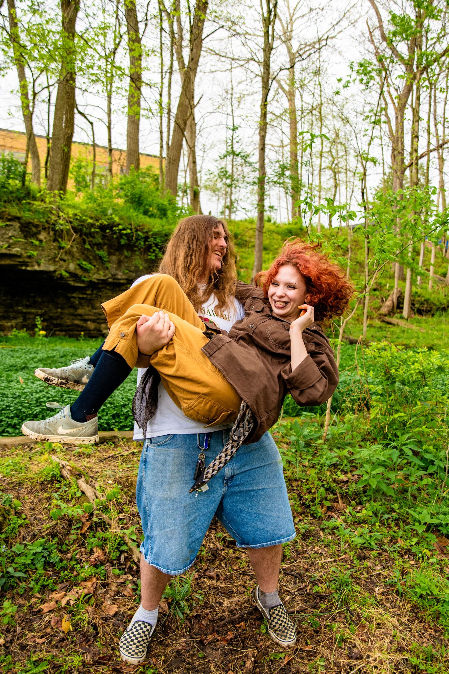 A woman with red curly hair carries her laughing friend piggyback style in a lush green forest setting.