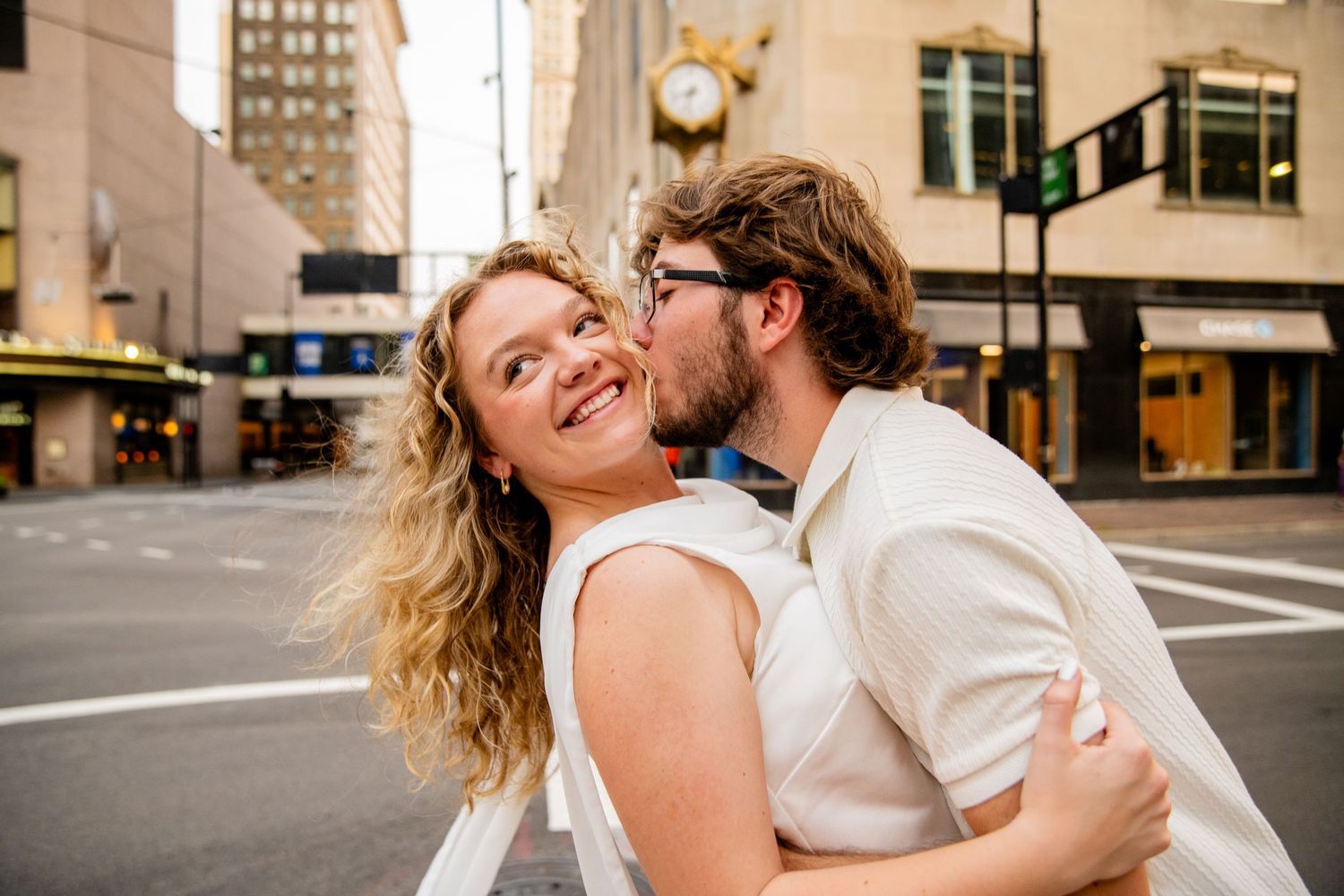 A couple shares a romantic kiss on a city street, the woman smiling joyfully in a white dress.