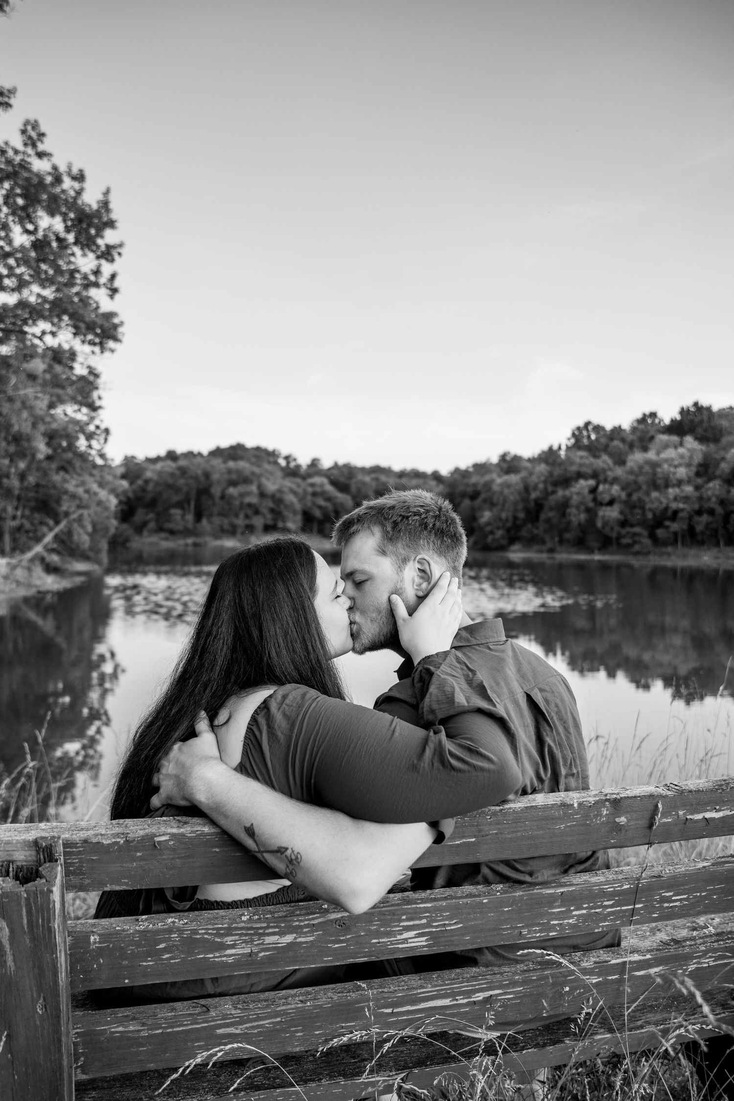 Black and white photo of a couple kissing on a wooden bench by a serene lake surrounded by trees.