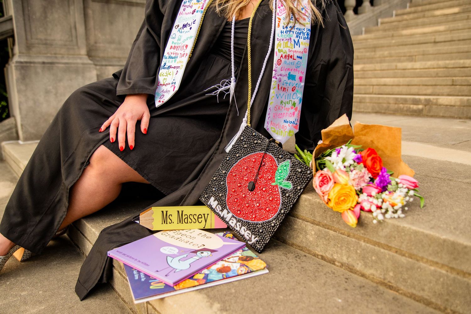 Graduate in black cap and gown with colorful stole, decorated mortarboard reading Ms. Massey, beside a floral bouquet.