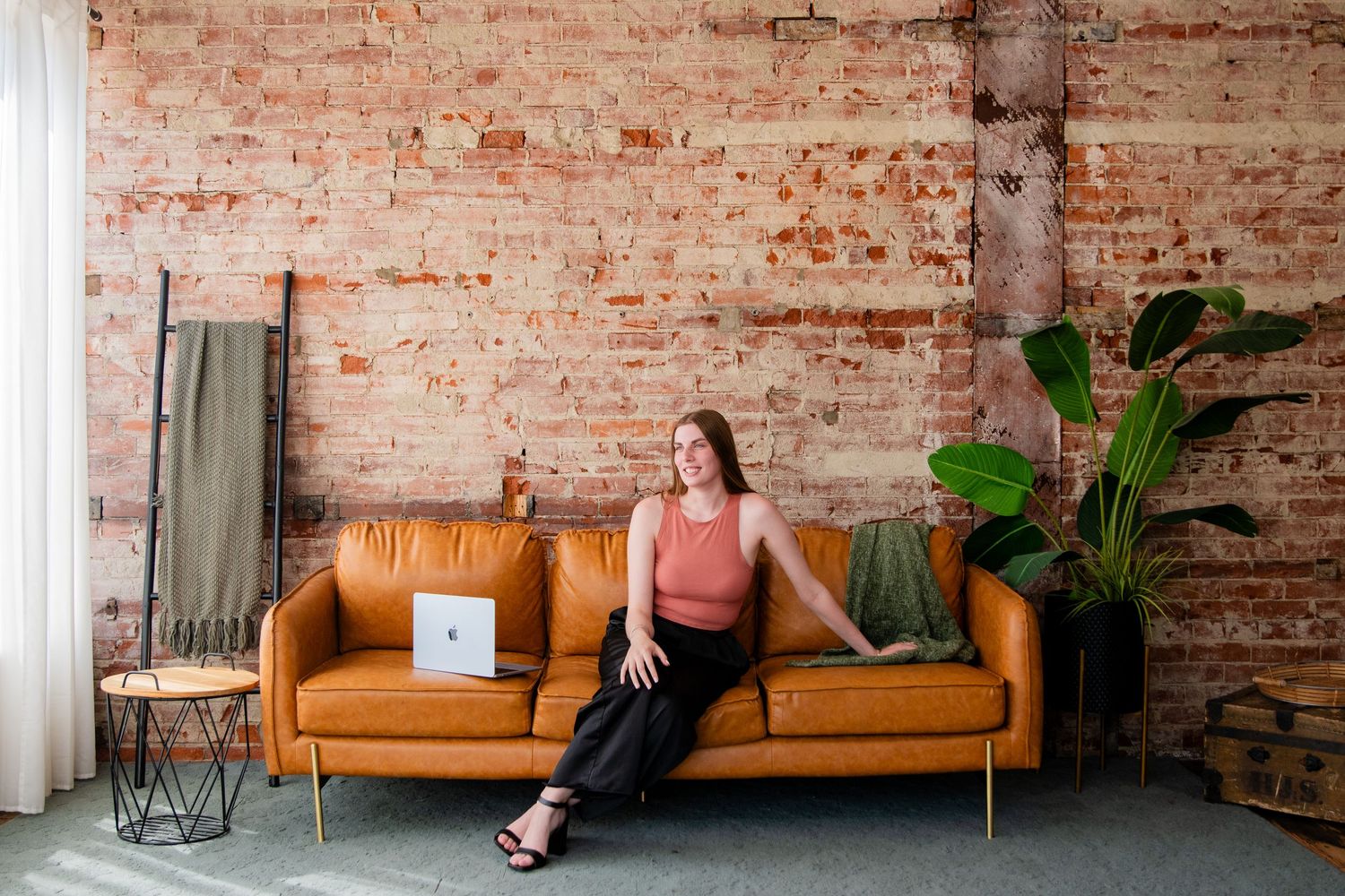 Woman relaxing on tan leather sofa with laptop, brick wall background, indoor plant, modern loft interior.
