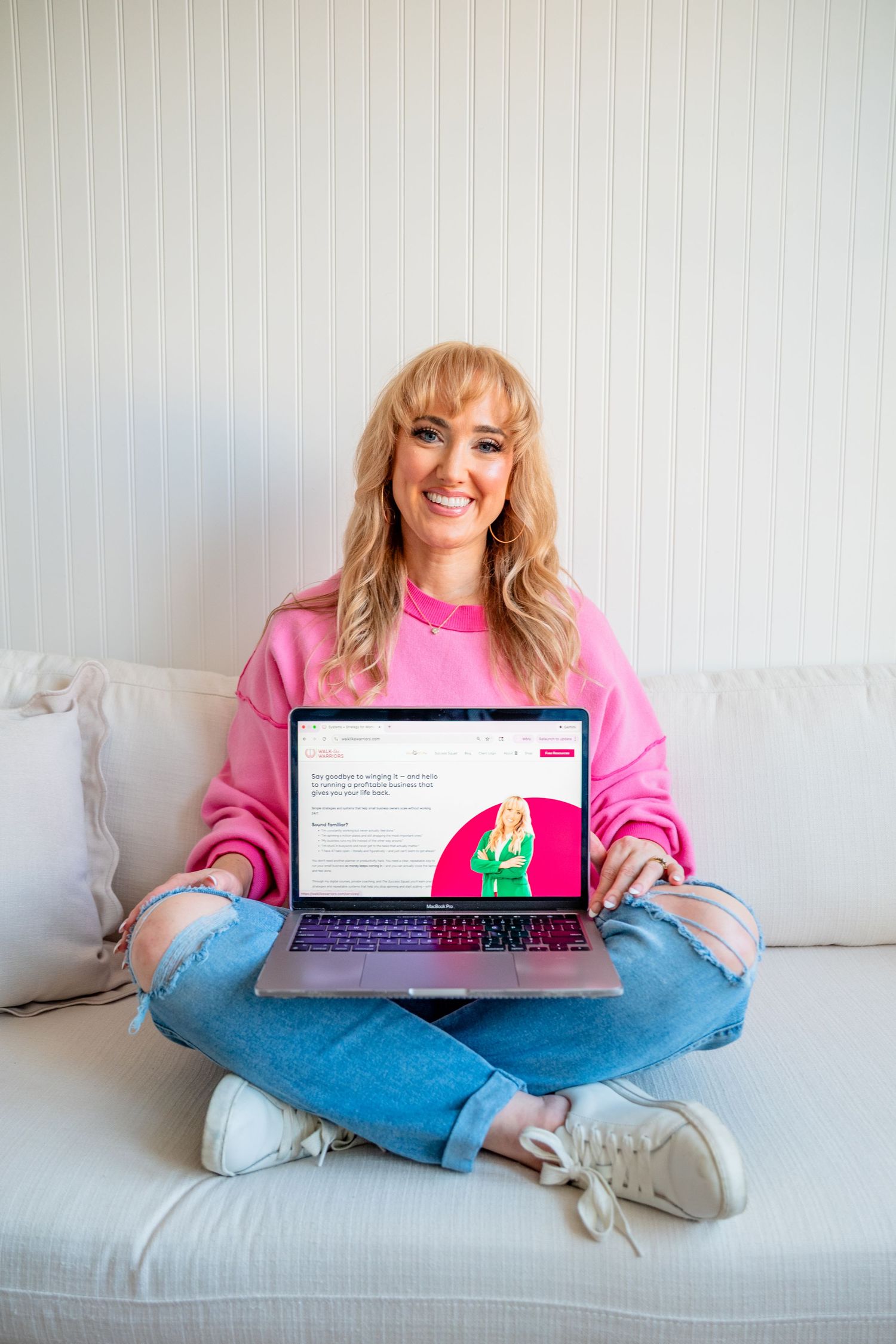 Smiling blonde woman in pink sweatshirt sitting cross-legged on sofa holding laptop showing website.
