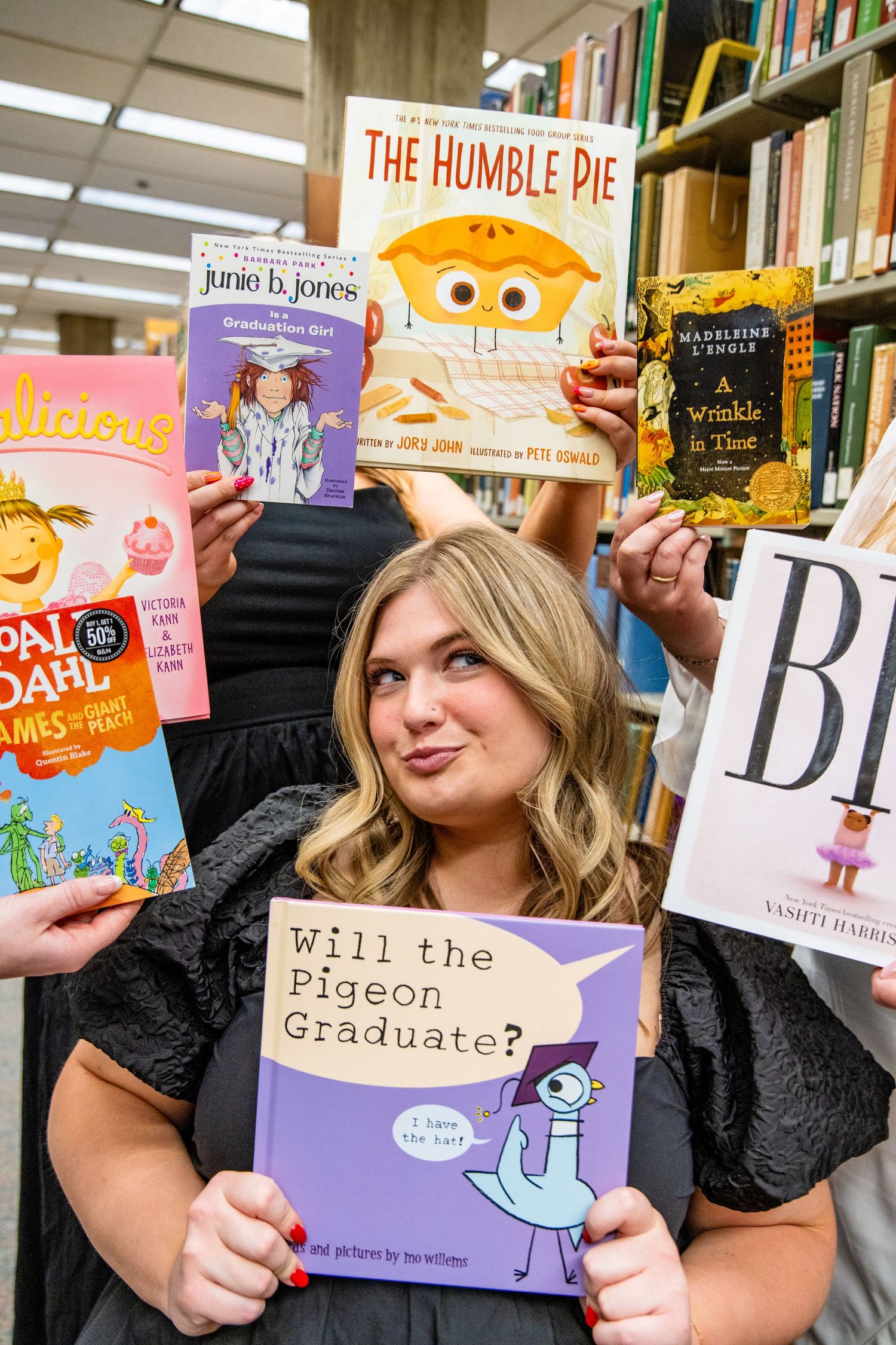Graduate poses with books including Will the Pigeon Graduate and The Humble Pie in a bookstore celebration.