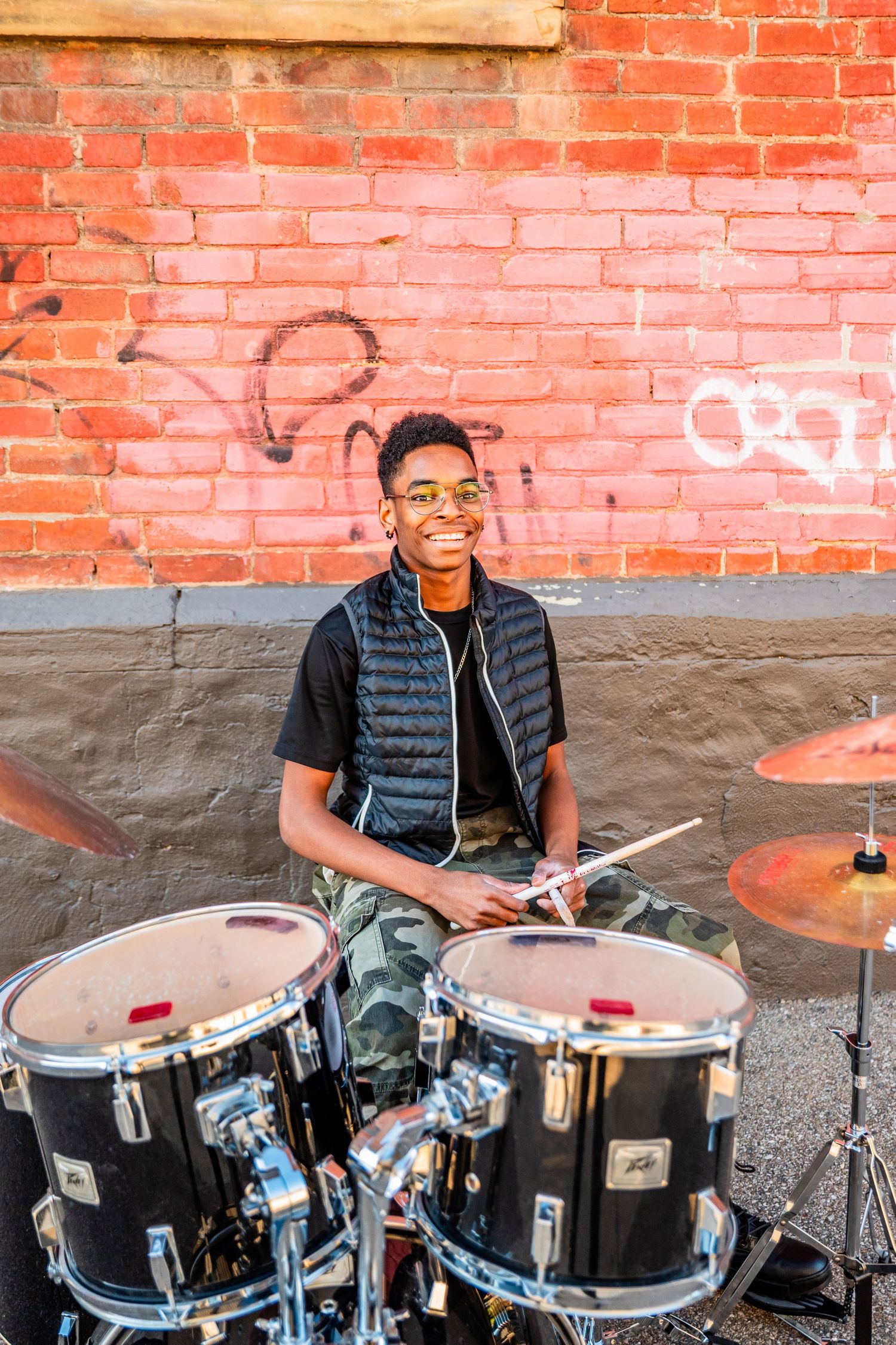 Smiling young Black drummer playing drums outdoors against a red brick wall with graffiti, wearing sunglasses and a vest.