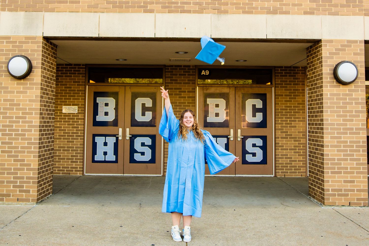 A smiling graduate in a blue cap and gown tosses her cap in the air outside BCHS high school entrance.