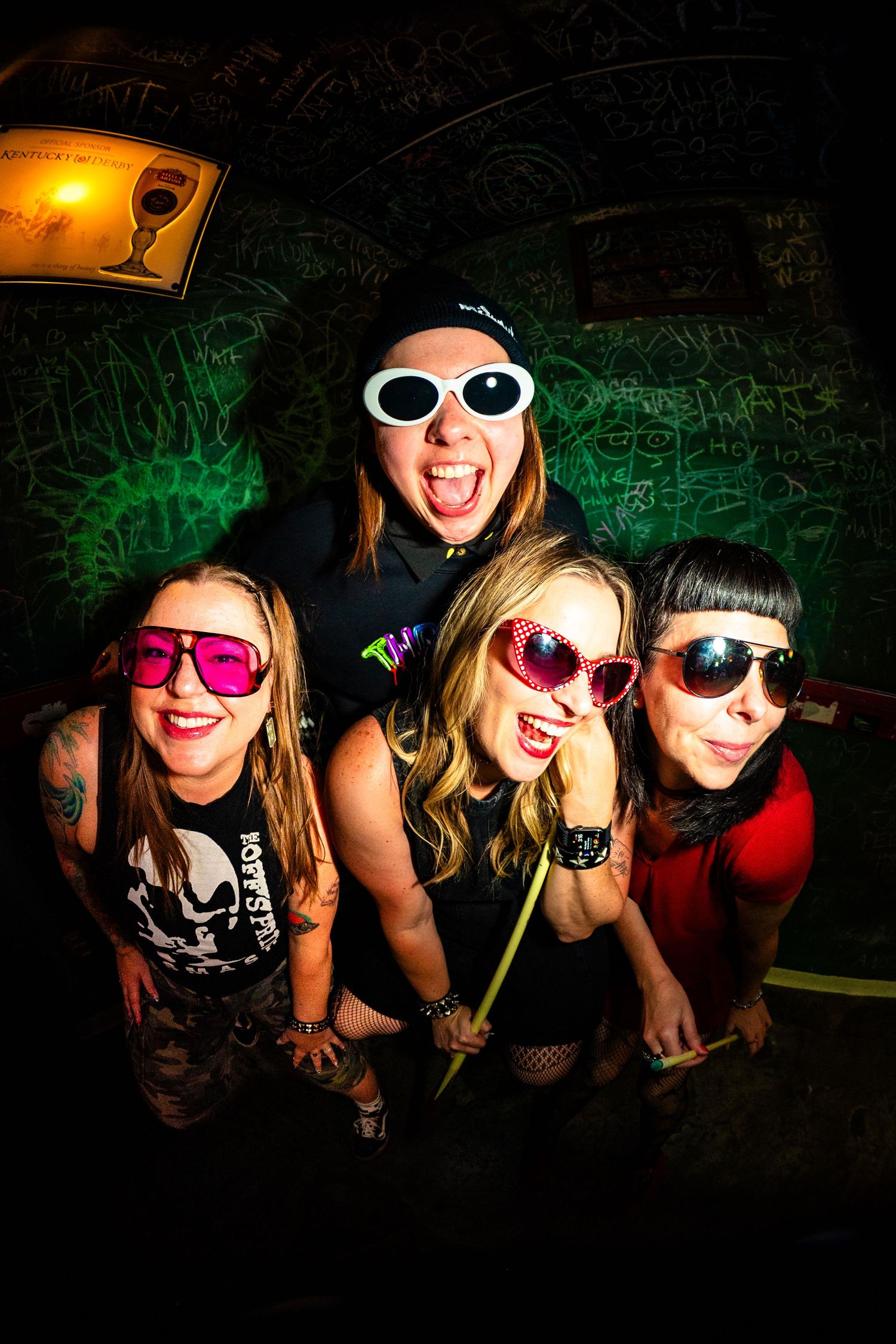 Four women in colorful funky sunglasses pose energetically in a graffiti-covered venue, exuding punk rock vibes.