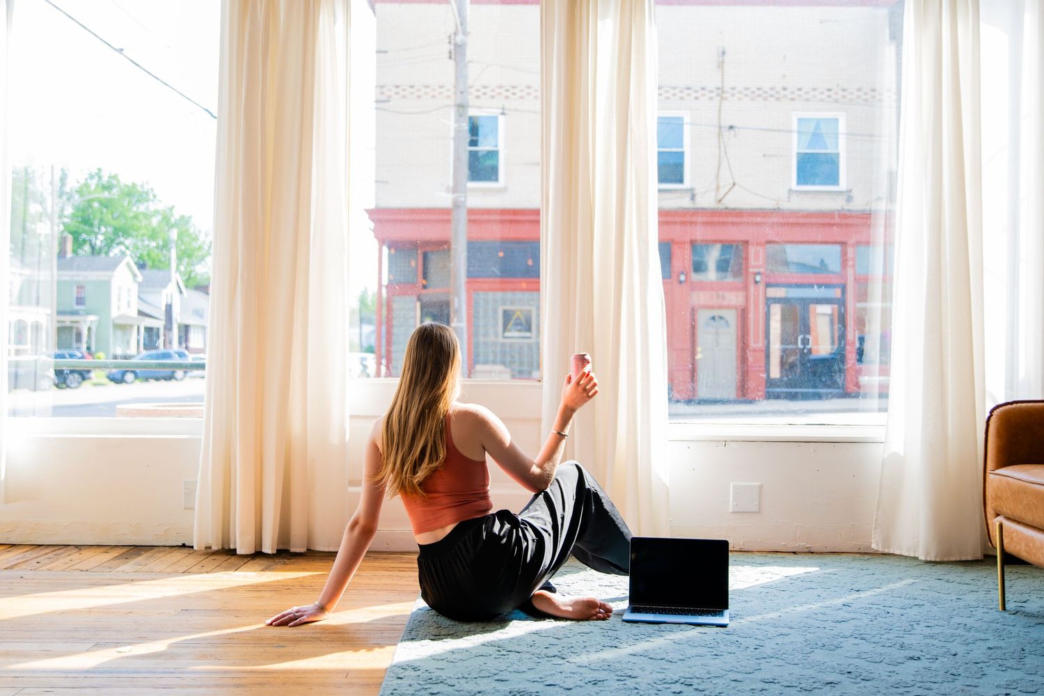 Woman sitting on yoga mat by large windows, stretching and relaxing in a bright, sunlit living room with hardwood floors.