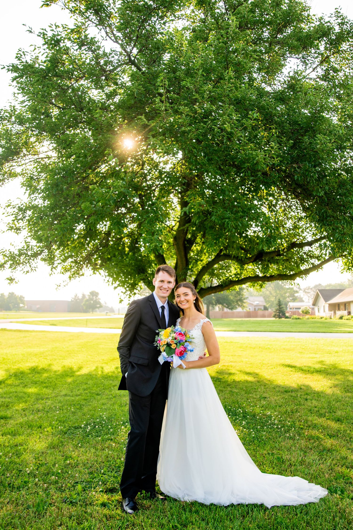 Happy bride and groom stand together under sunlit green tree, bride holding vibrant colorful wedding bouquet.