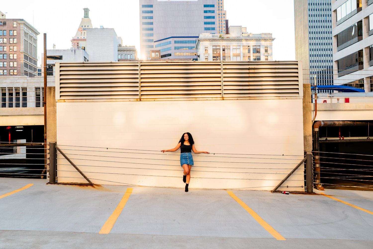 Young woman dancing on rooftop parking lot with urban city skyline buildings in background on sunny day.