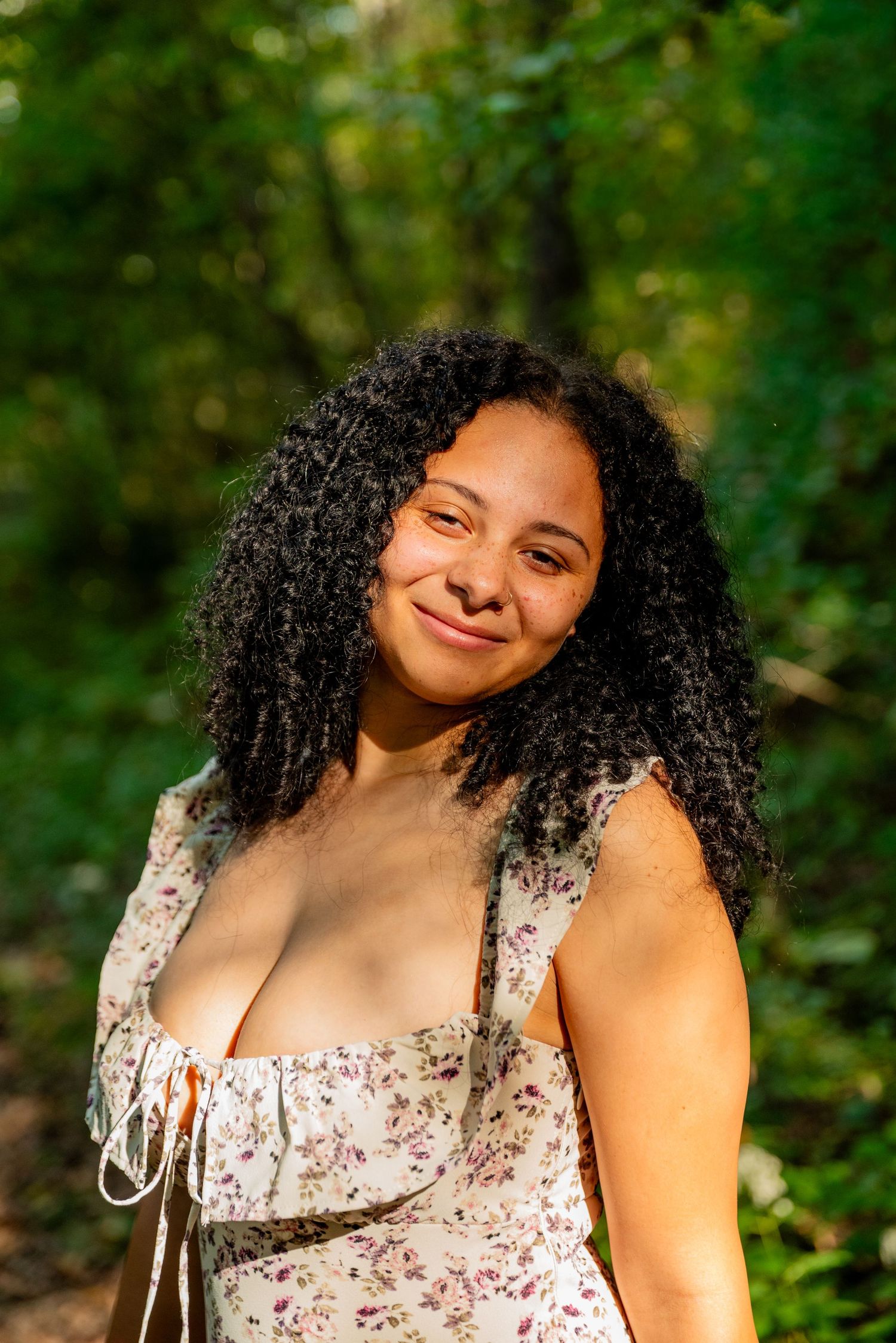 Young woman with curly black hair smiling in a floral dress outdoors in a sunlit forest setting.