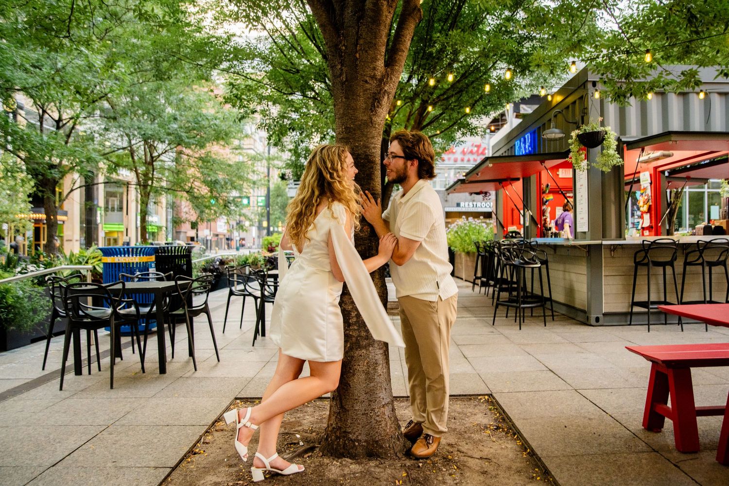Couple sharing a romantic moment by a tree on a lively urban patio with string lights and outdoor seating.