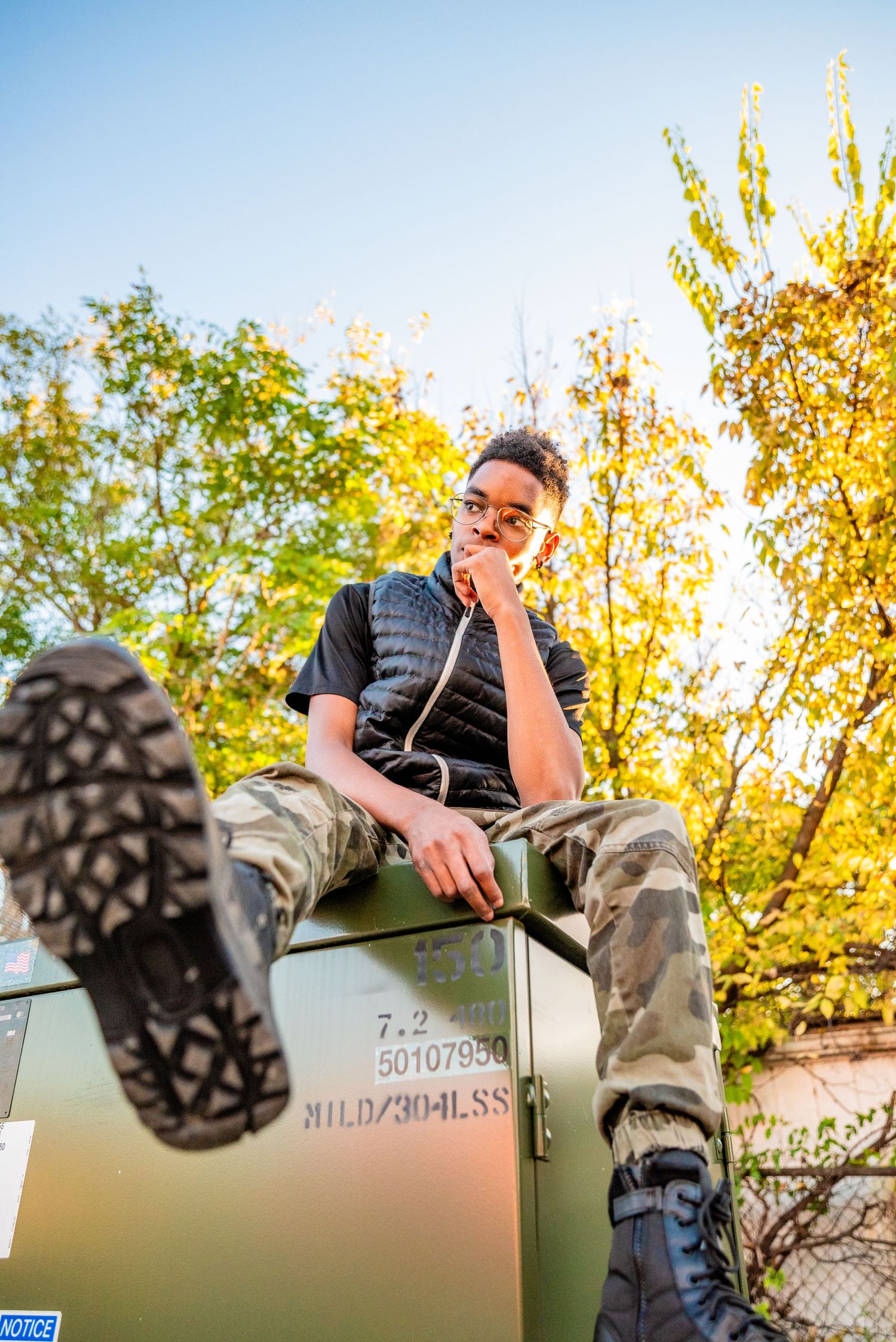 Young man in camo pants and black shirt sits atop a military green box outdoors, surrounded by autumn trees.