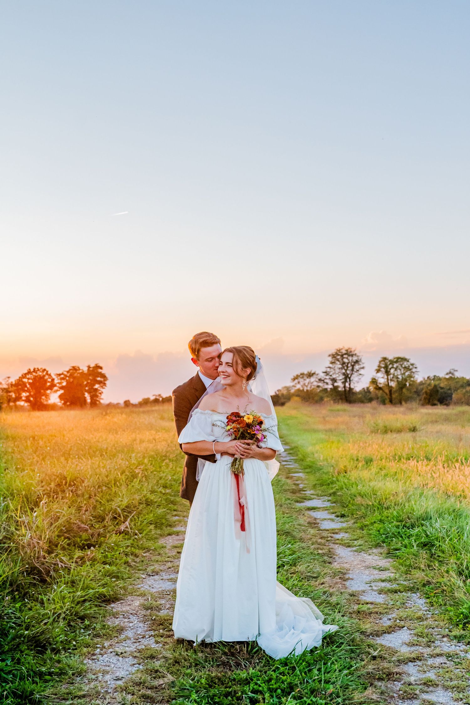 Bride and groom embrace on a rustic dirt path in a golden sunset field, bride holding autumn bouquet.