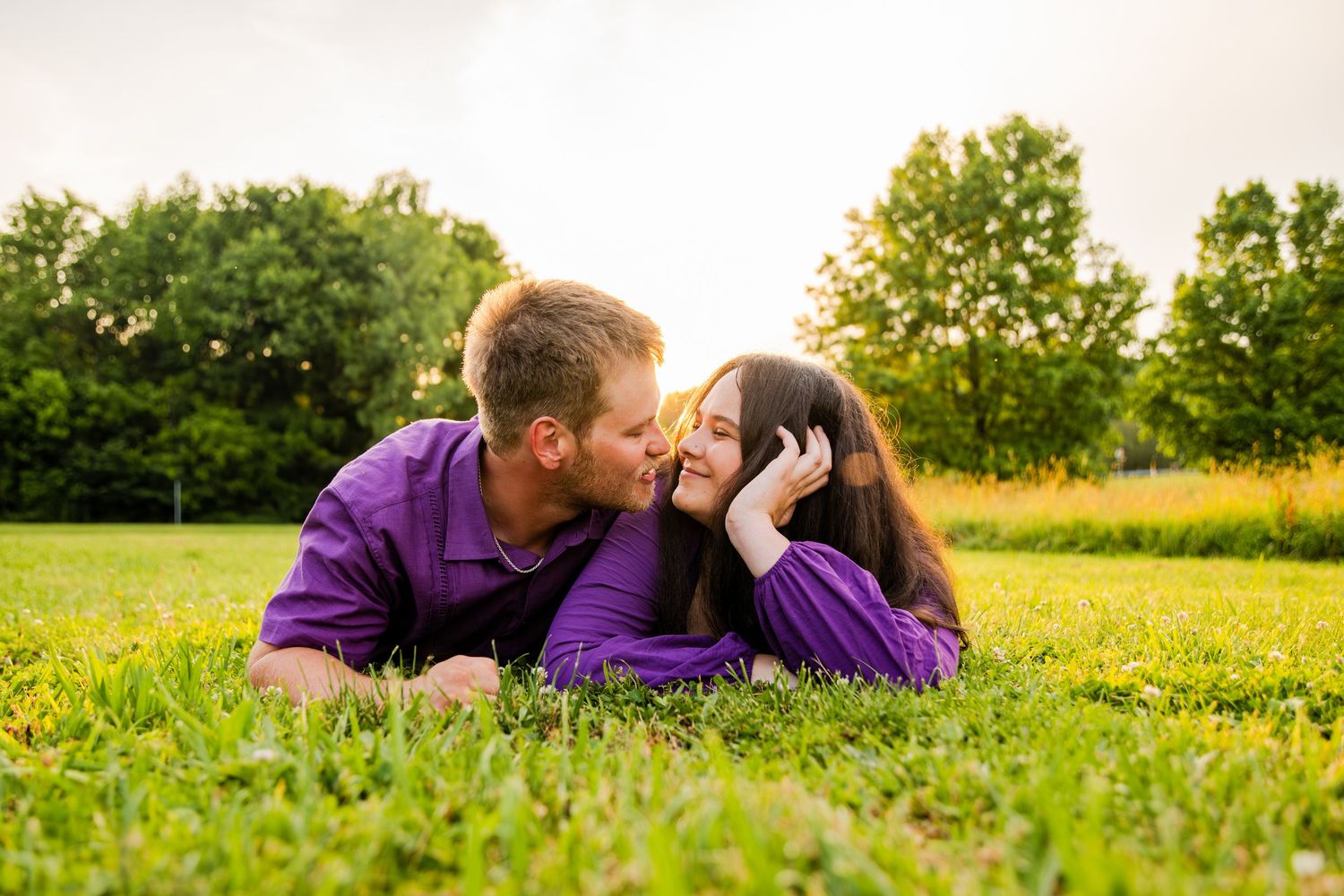 Couple in matching purple shirts lying on green grass, sharing a romantic kiss outdoors with trees in background.