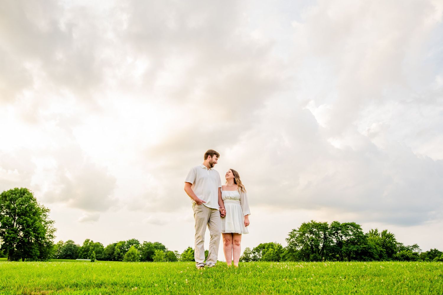 Couple holding hands in a lush green field under dramatic cloudy sky, sharing a romantic outdoor moment.